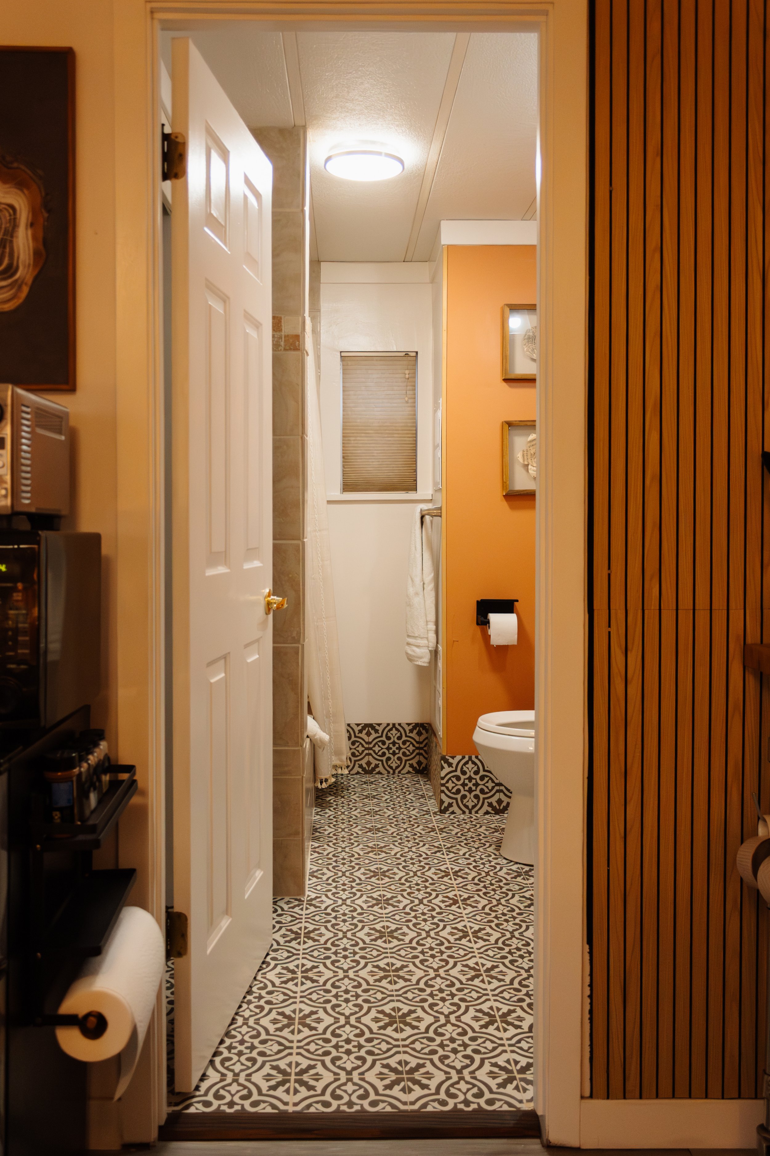 View of a compact bathroom with a patterned floor, white toilet, orange accent wall, small window with blinds, and a shower with a curtain.