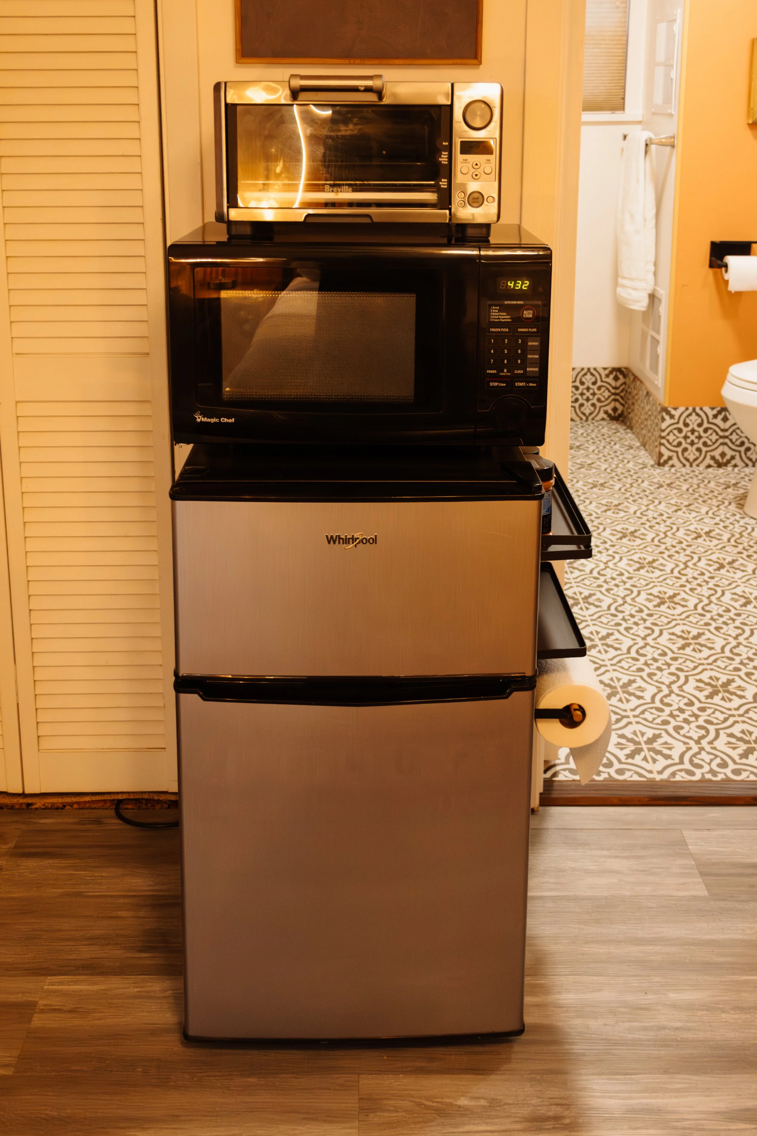 A small kitchen area with a stainless steel mini fridge, a black microwave oven, and a stainless steel toaster oven stacked on top of each other. The floor is wood and there is a roll of toilet paper hanging on the side of the mini fridge.