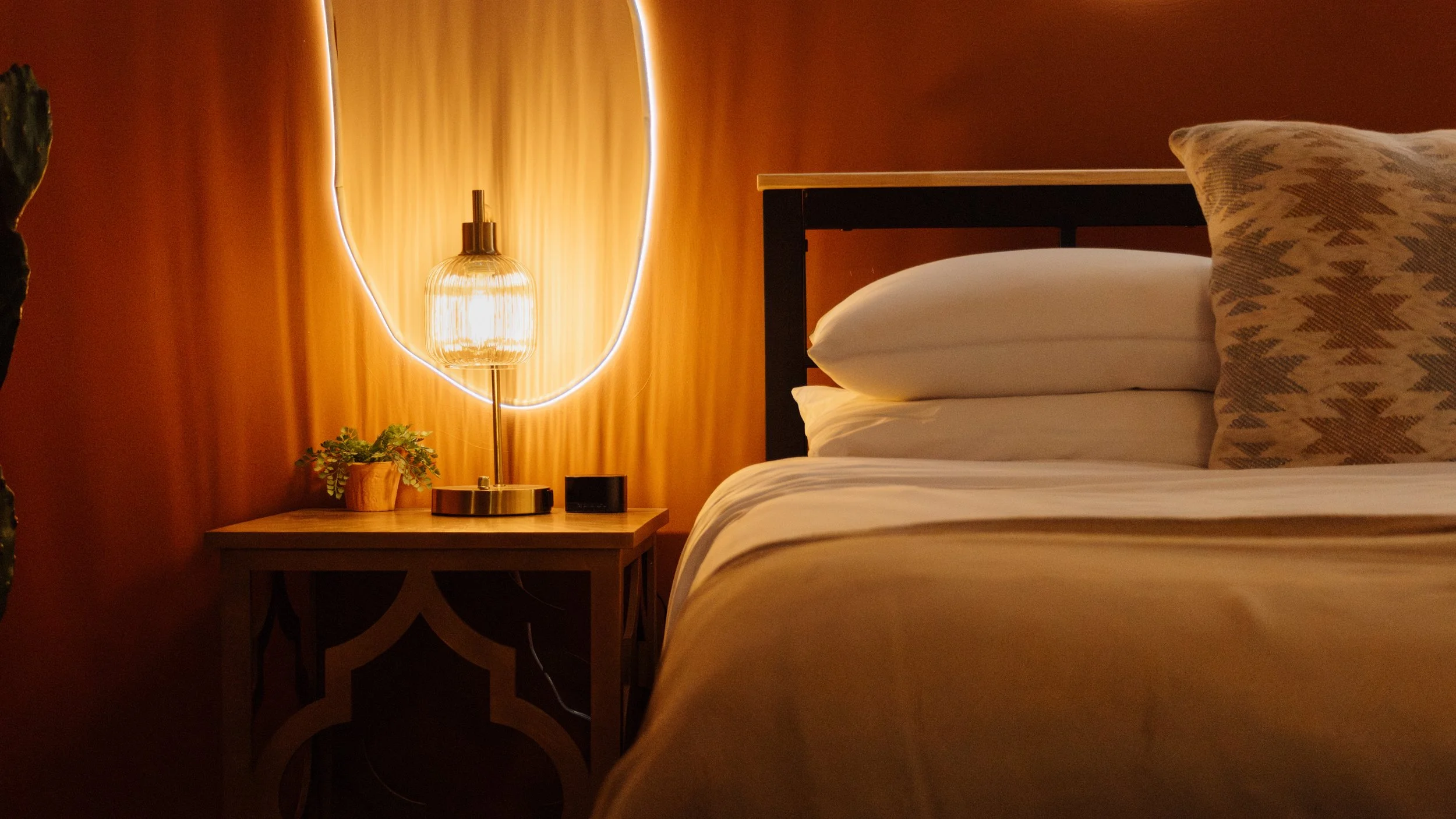 A cozy bedroom corner with a bed featuring white linens and a decorative patterned pillow. A wooden nightstand holds a modern table lamp with a golden base and warm light, along with a small potted plant and a black alarm clock. The background has a 