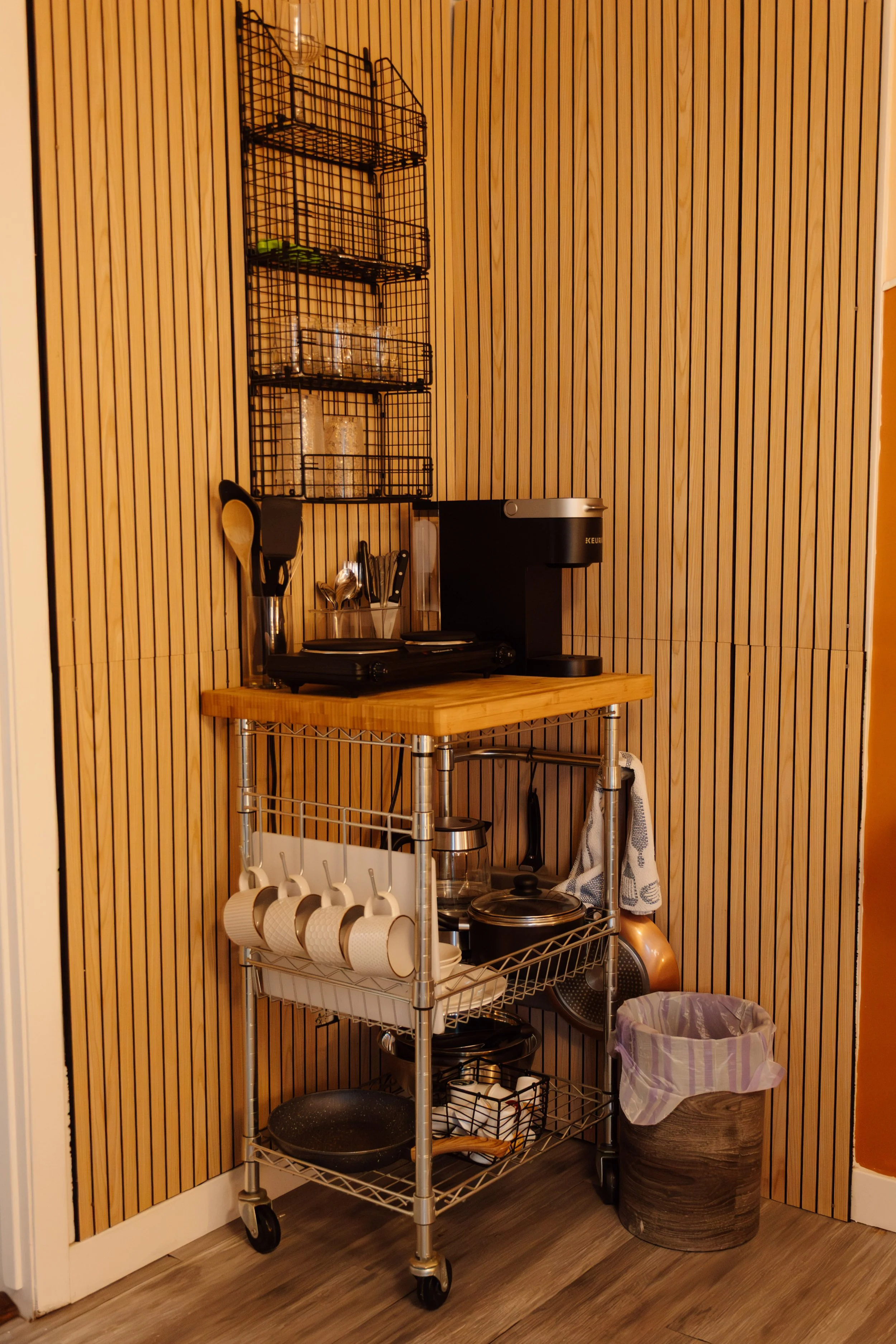 A kitchen cart with a wooden top, holding a coffee machine and utensils, with shelves below containing cups, a pan, and miscellaneous kitchen items, adjacent to a wooden wall and trash can.