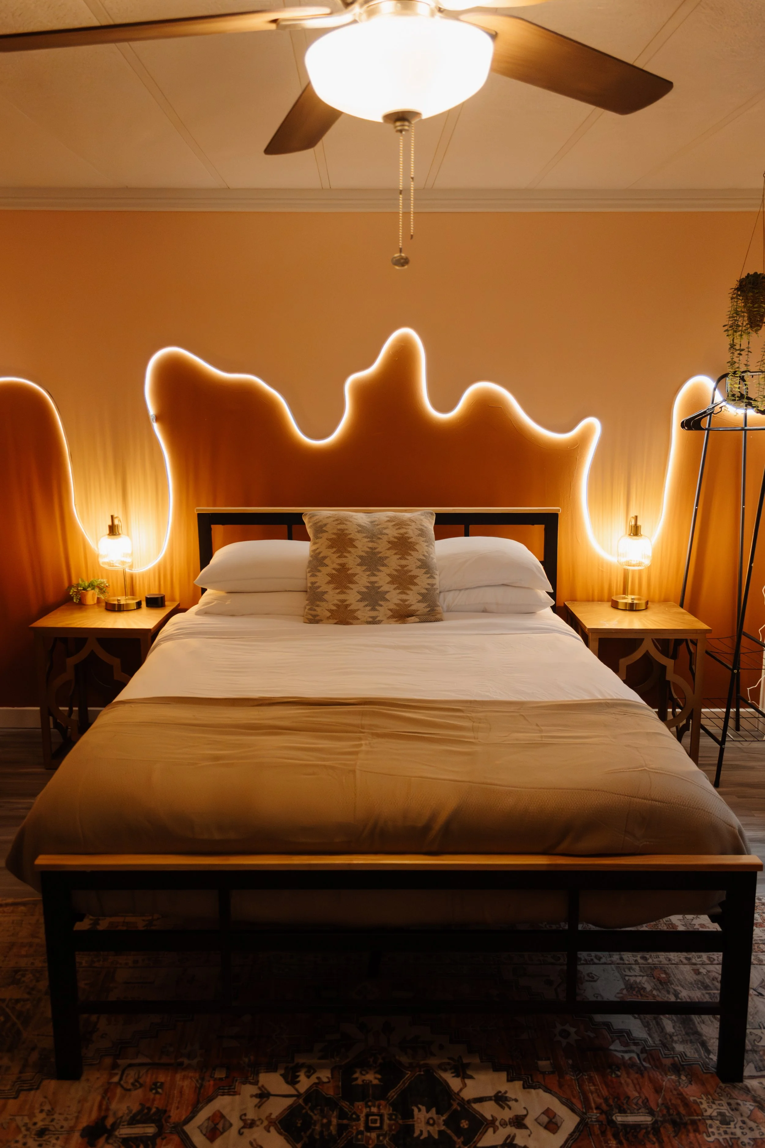 A cozy bedroom featuring a bed with a beige bedspread, striped pillow, and two white pillows. The headboard has a wavy design outlined with neon lighting, and matching bedside tables with lamps. A ceiling fan is mounted on the ceiling.