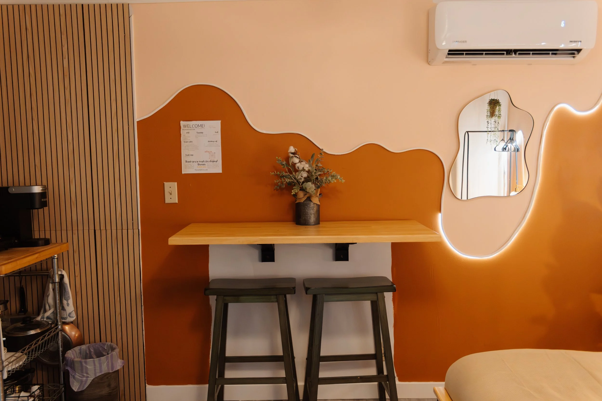 Decorative interior wall with wavy two-tone paint in warm colors, small mirror, air conditioner, and a wooden table with a flower arrangement. Seating with two stools and kitchen items on a cart.