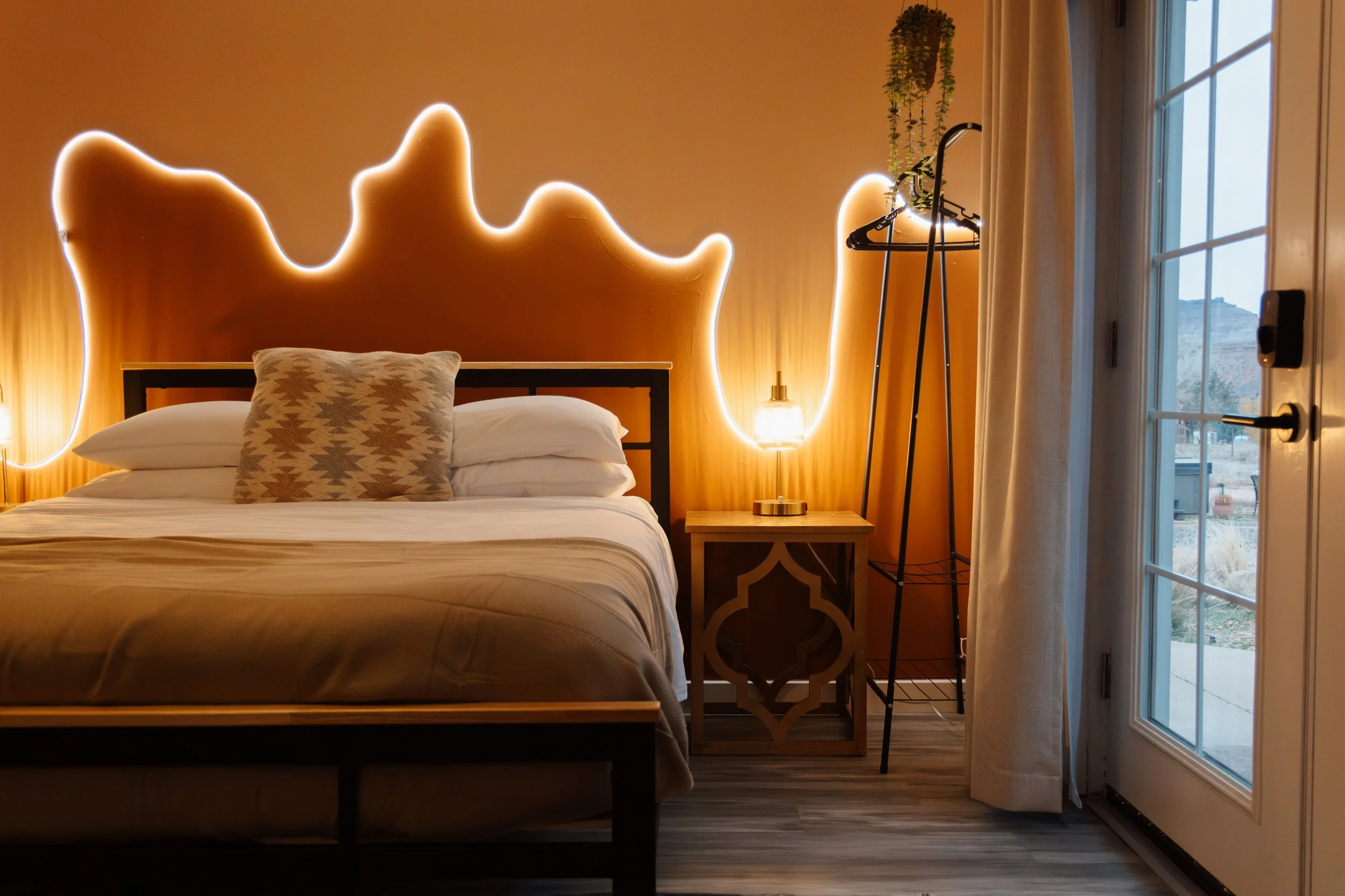 Cozy bedroom with a wooden bed, patterned pillow, a nightstand with a table lamp, a hanging plant, a clothing rack, and a glass door with curtains revealing an outdoor landscape.