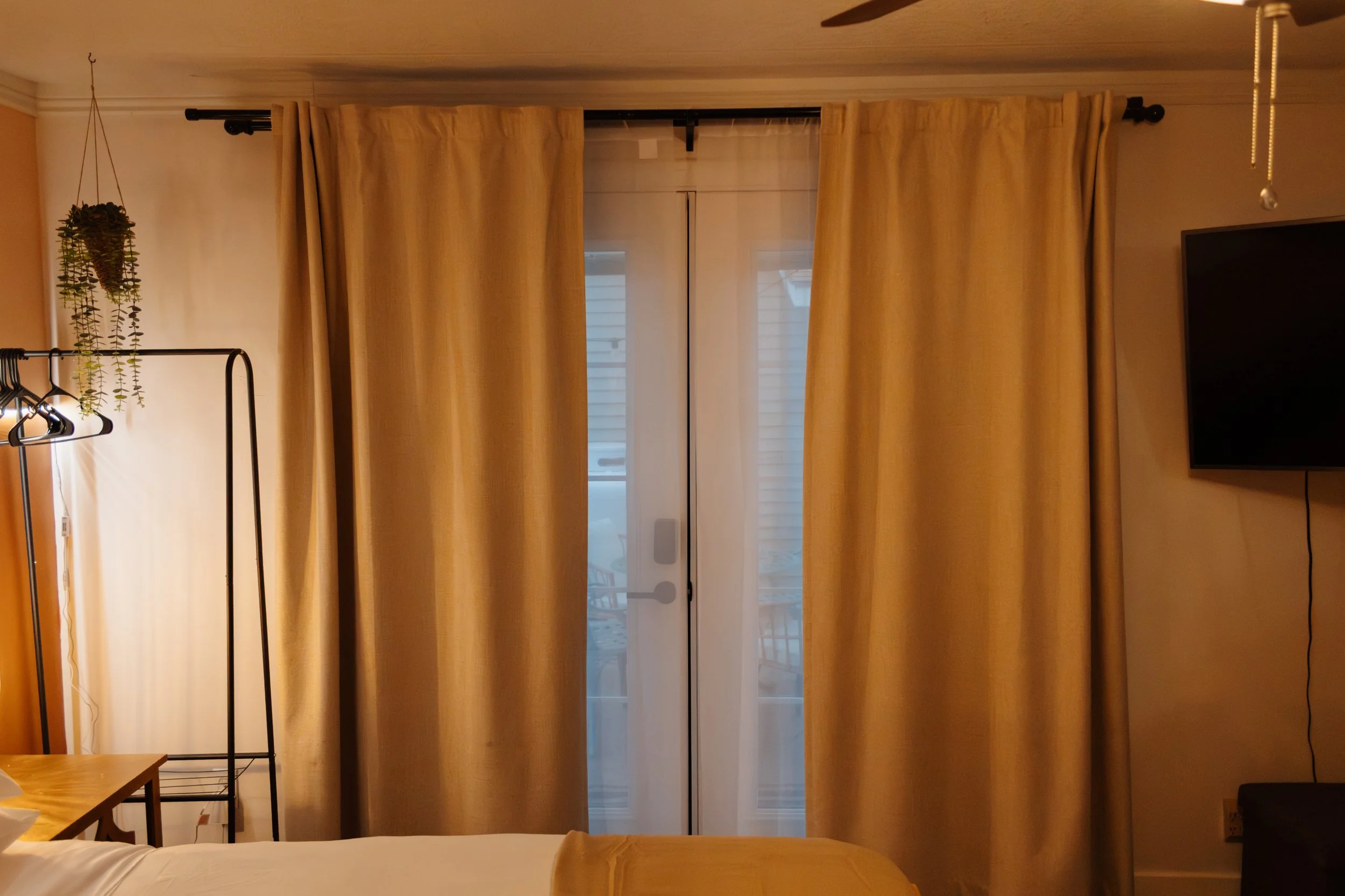 A room with beige curtains covering a glass door leading to a balcony, a corner with a hanging plant and a coat rack, and a wall-mounted flat-screen TV on the right.