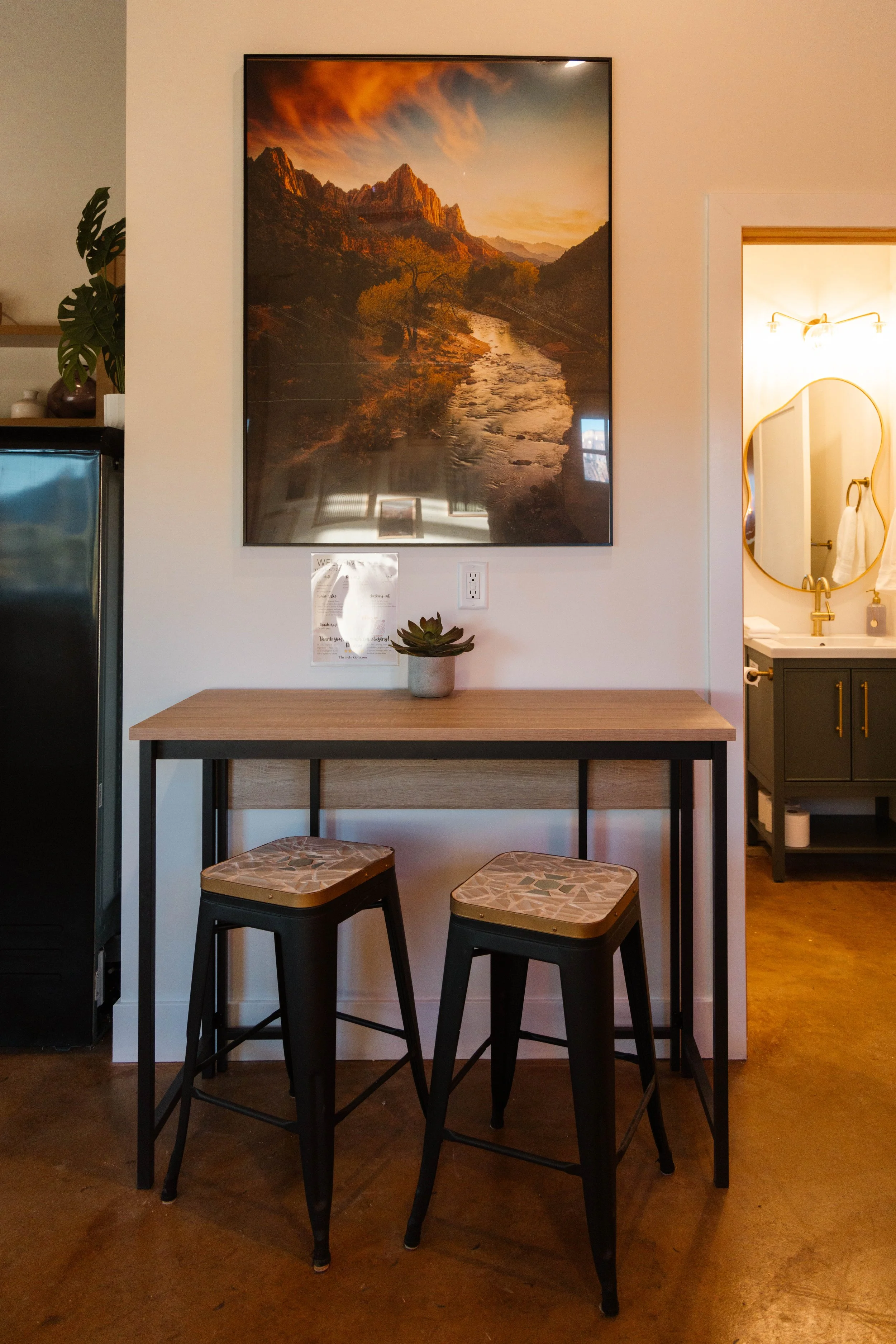 A small wooden table with two stools featuring mosaic tops, placed against a white wall. Above the table, a large framed landscape photograph of mountains, river, and sunset or sunrise. A small potted succulent sits on the table, with a paper sign and a wall outlet nearby. To the right, part of a bathroom mirror and sink are visible.