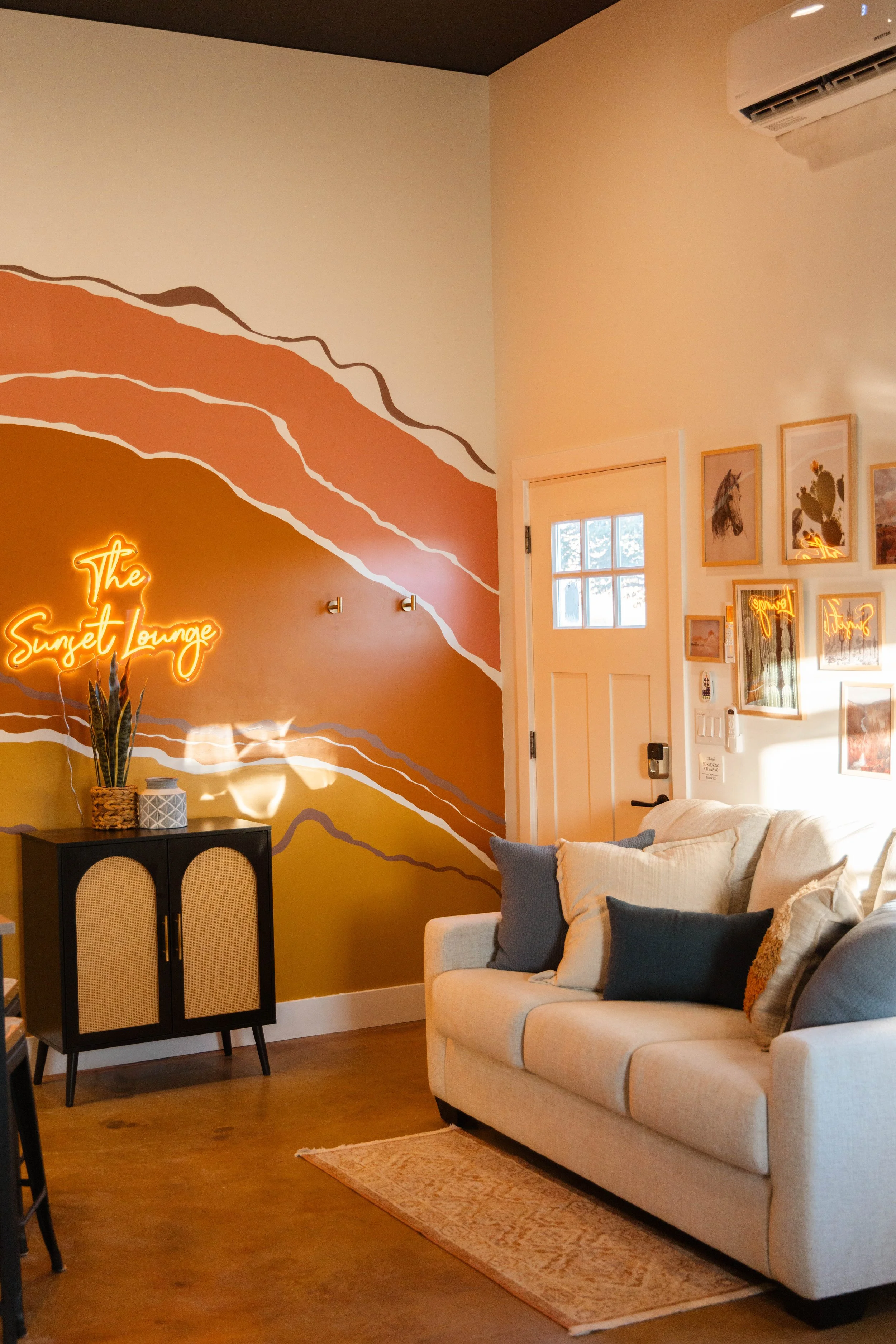 Living room with a cream sofa and stacked pillows, wall art, a black cabinet with potted plant, and a neon sign that says "The Sunset Lounge" on a colorful mural wall.