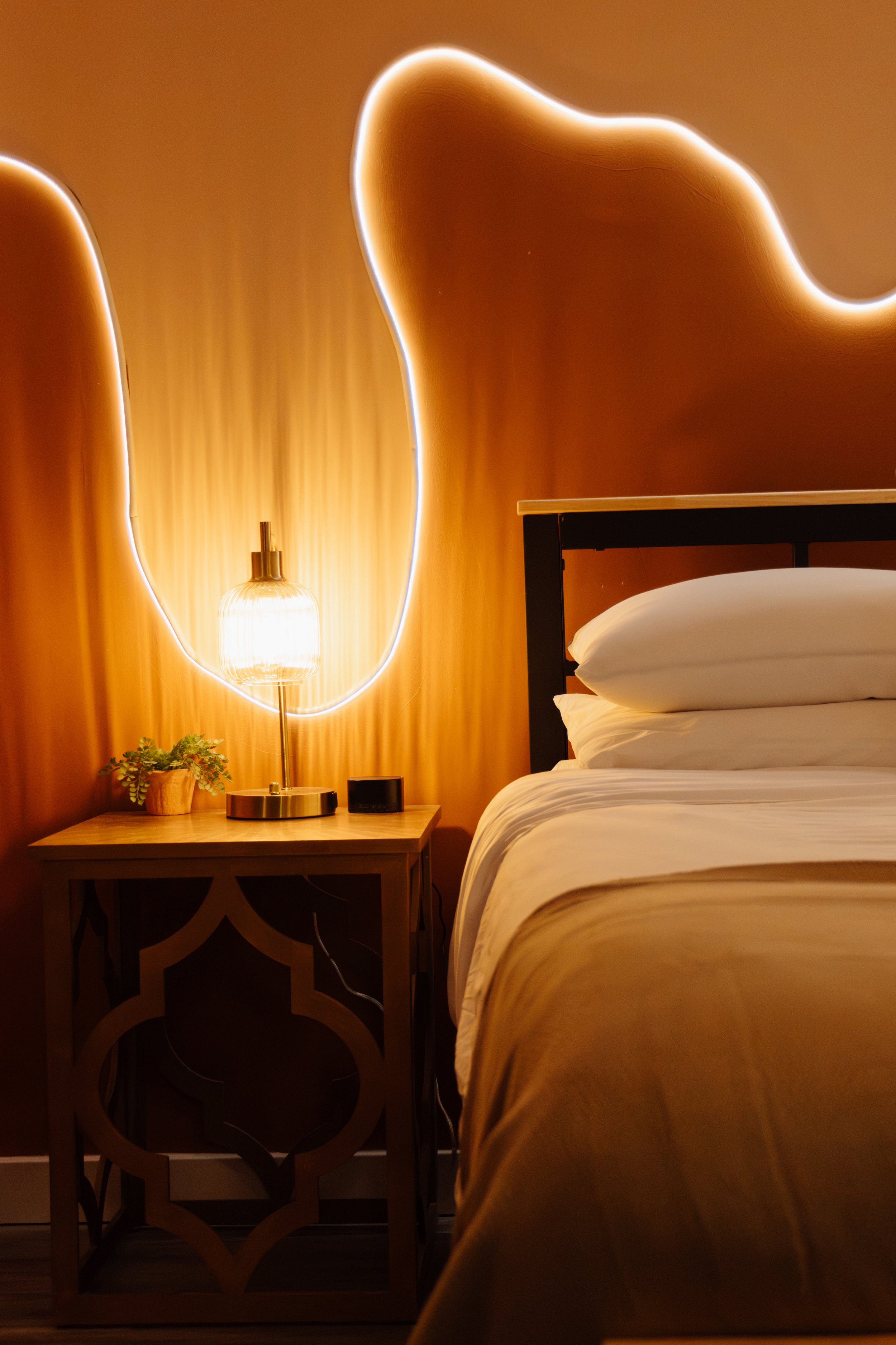A bedroom with a bed, white pillows, a wooden nightstand, a lamp, a small potted plant, and a neon light art on the wall.