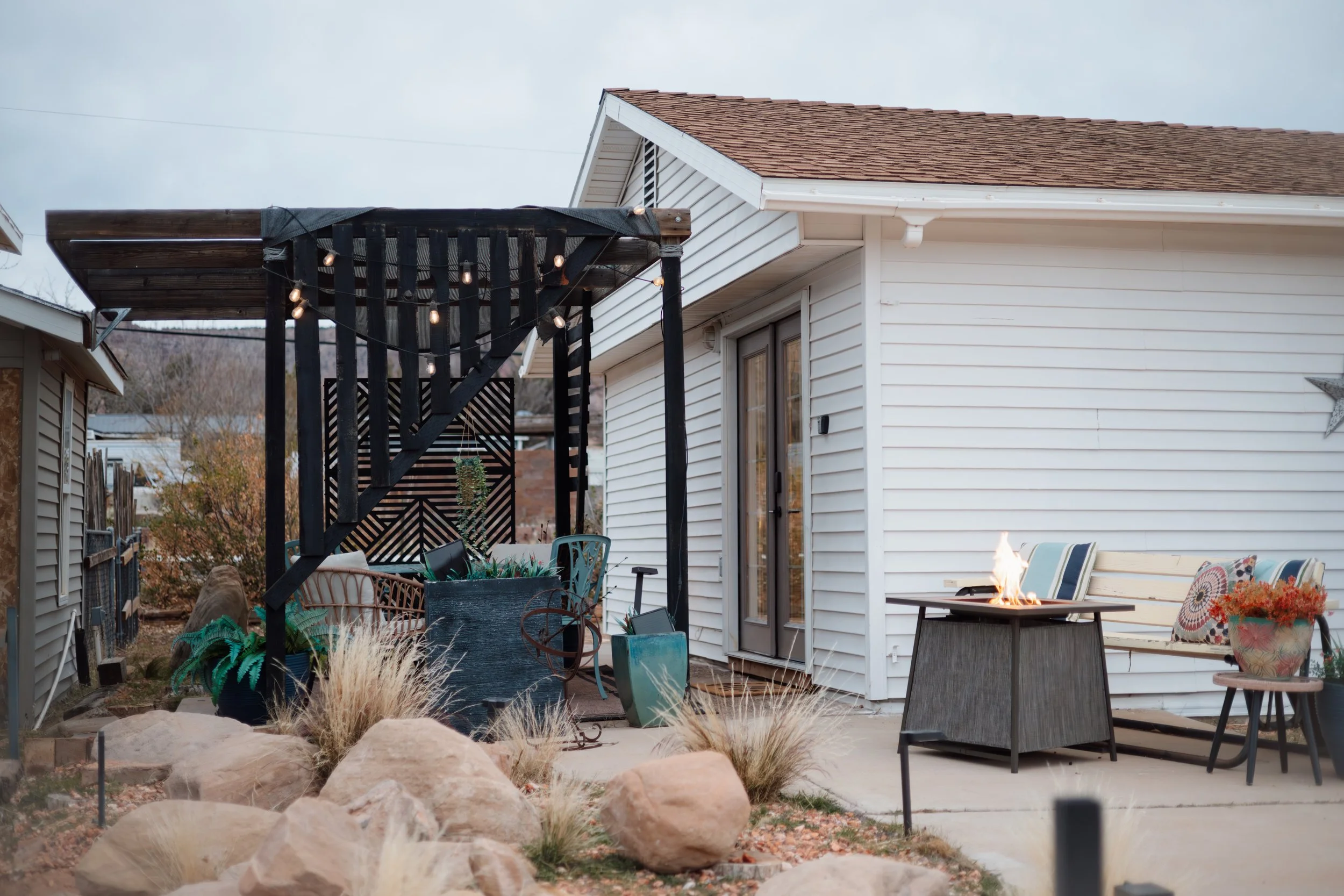 Backyard patio with a small fire table, white house with sliding glass door, decorative pillows, and string lights on a black wooden pergola.