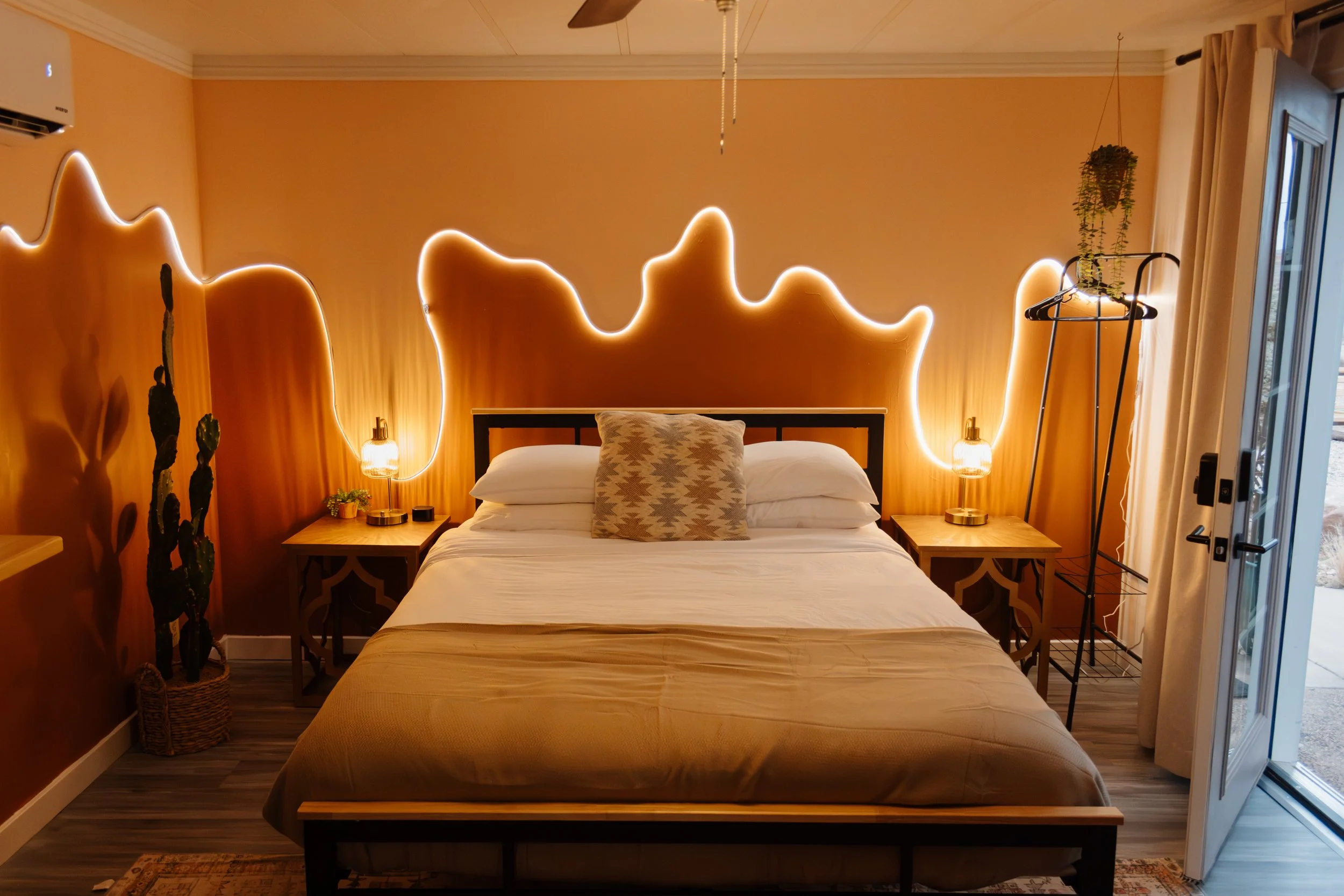 A bedroom with a bed, nightstands, and decorative lighting. The wall behind the bed has an artistic, wavy light strip. There are two small lamps on the nightstands, a plant in a basket on the left, and a hanging plant on the right near the door. The 