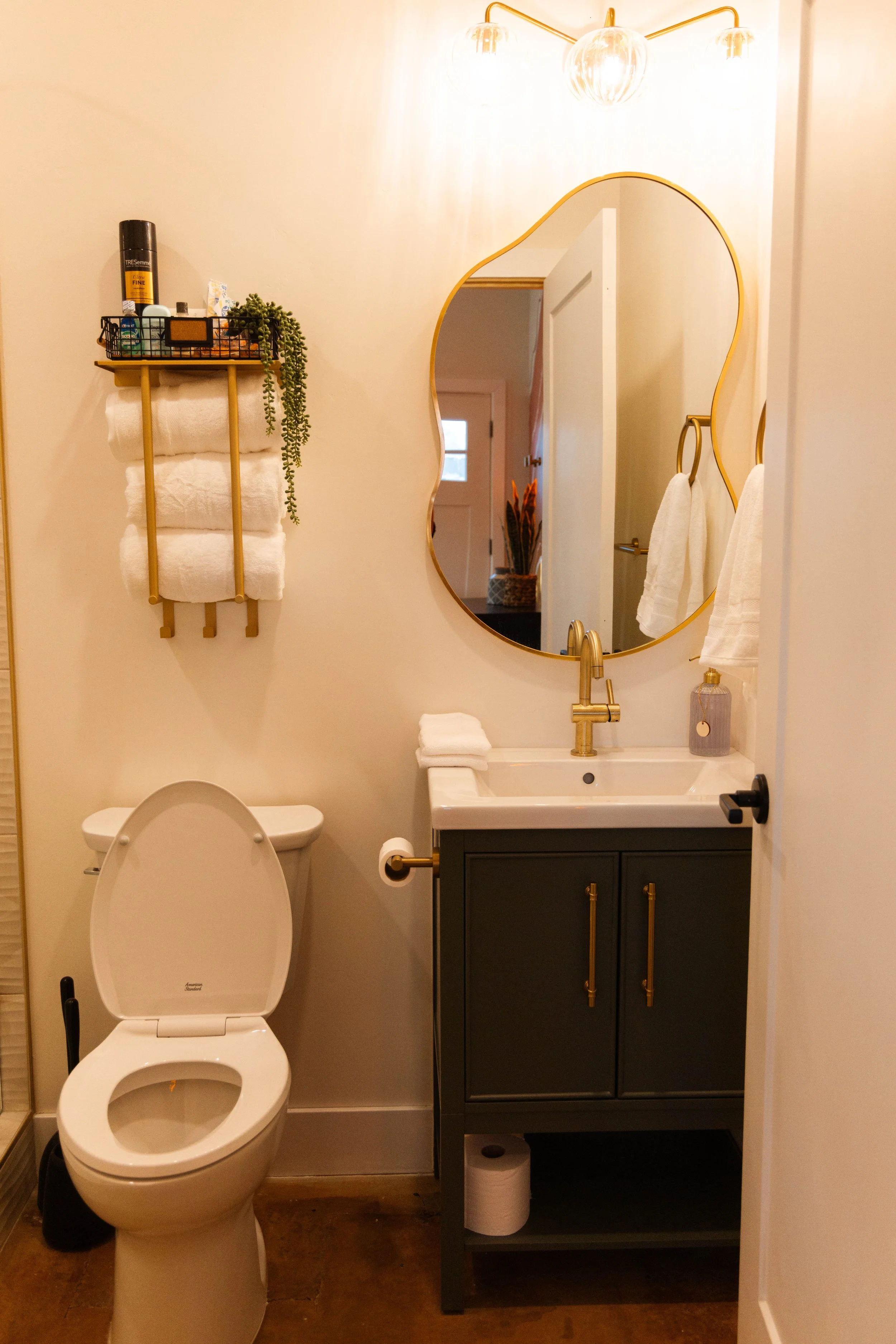 A small bathroom with a toilet, a vanity with a sink, a mirror, and wall-mounted towel rings holding white towels. There is a wooden shelf above the toilet holding rolled towels, toiletries, and a small plant. The bathroom has a gold and black tone theme with white walls and a wooden floor.