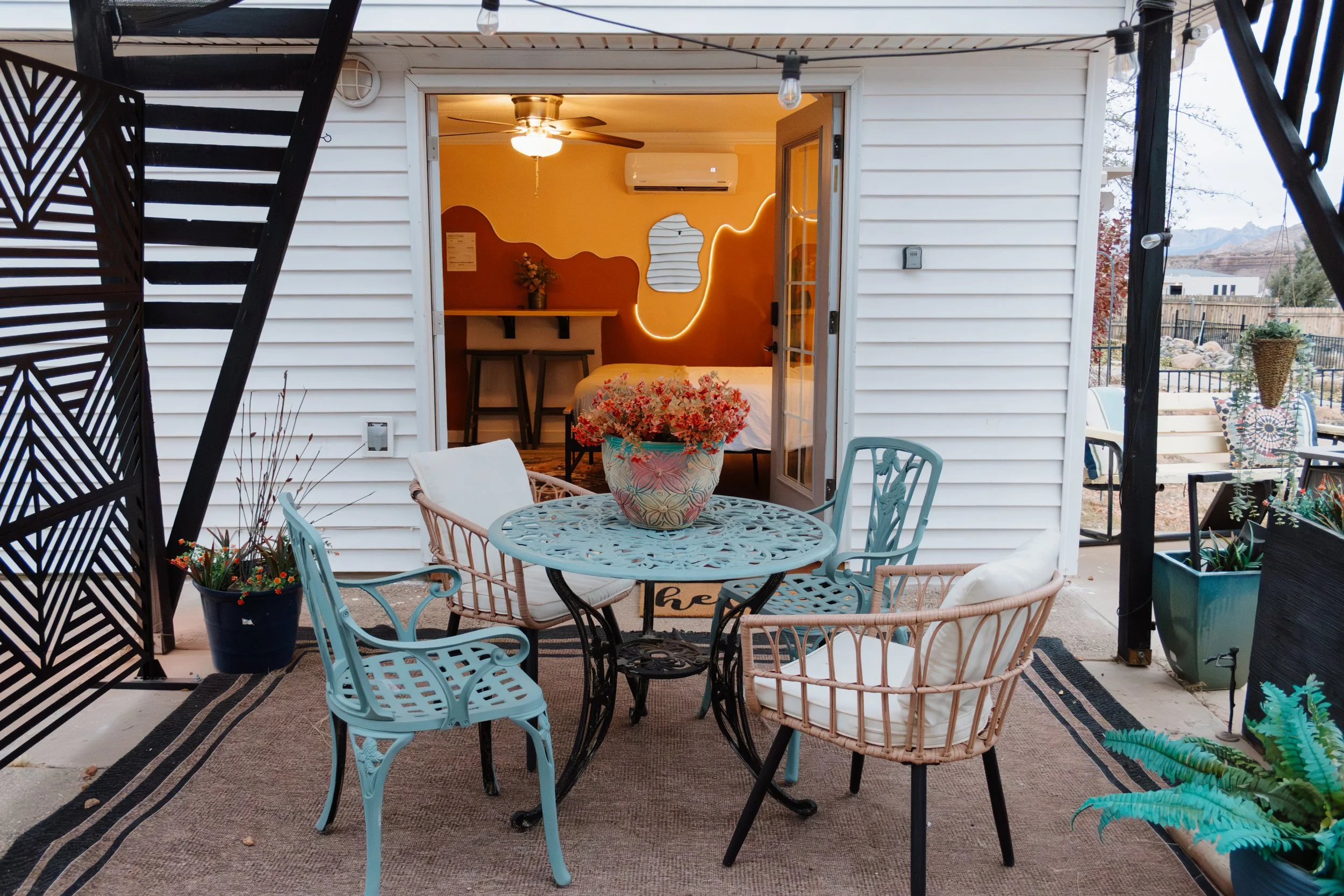 Outdoor patio with a round metal table and six mixed chairs, potted plants, and an open sliding door leading into a cozy bedroom with warm lighting, wall-mounted mirror, and air conditioning.