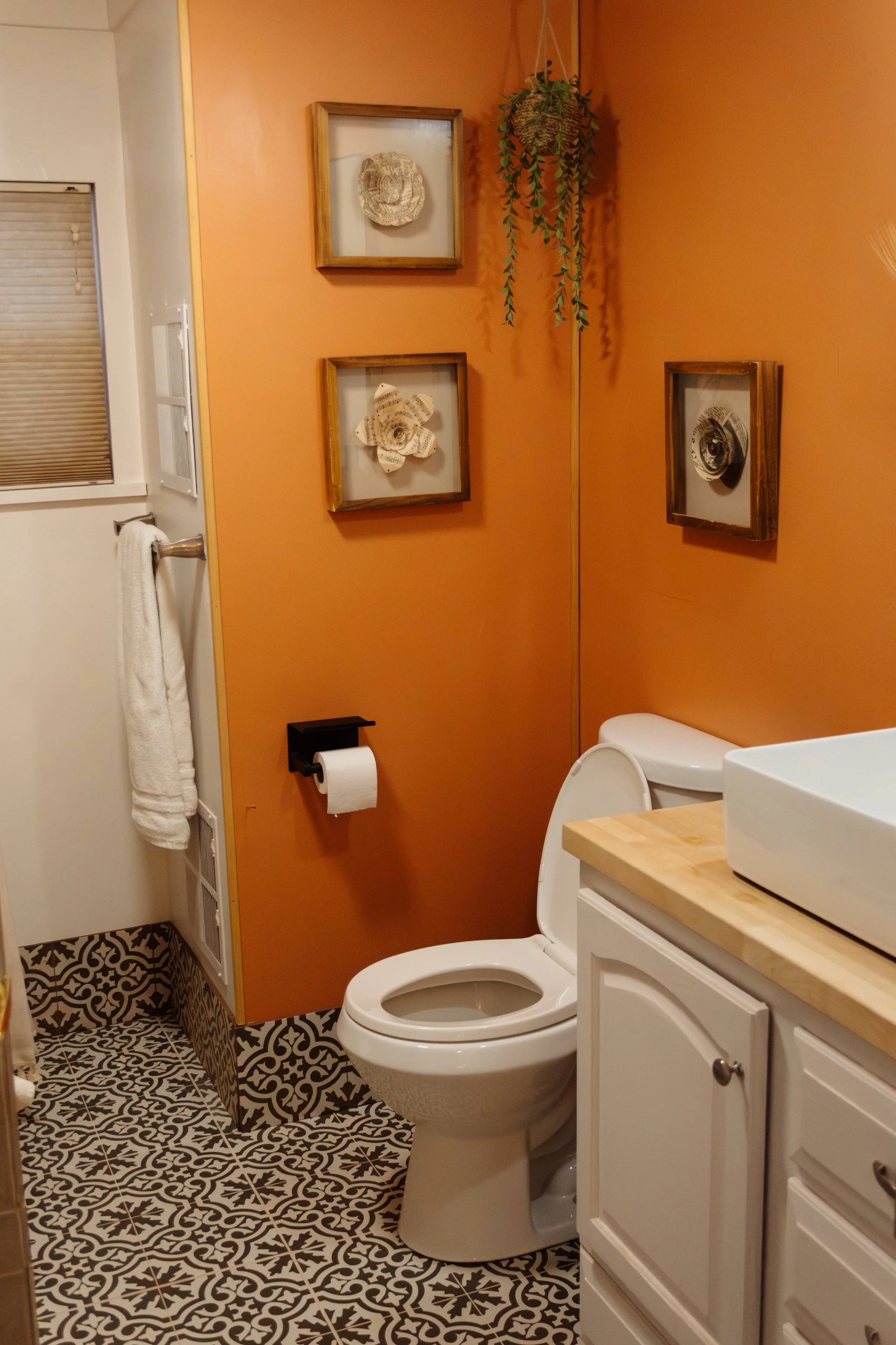 A small bathroom with orange wall paint, decorated with framed dried flowers. Contains a toilet, a small white vanity with a sink, a toilet paper holder, a towel rack with a white towel, and patterned black and white floor tiles.