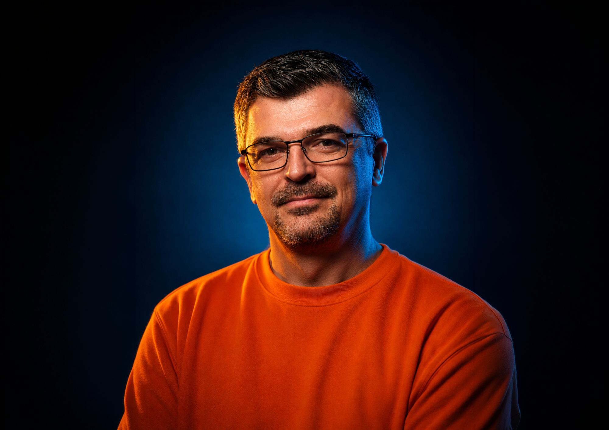 Portrait d'un homme portant des lunettes et un t-shirt orange, avec un fond sombre et un éclairage doux sur le visage.