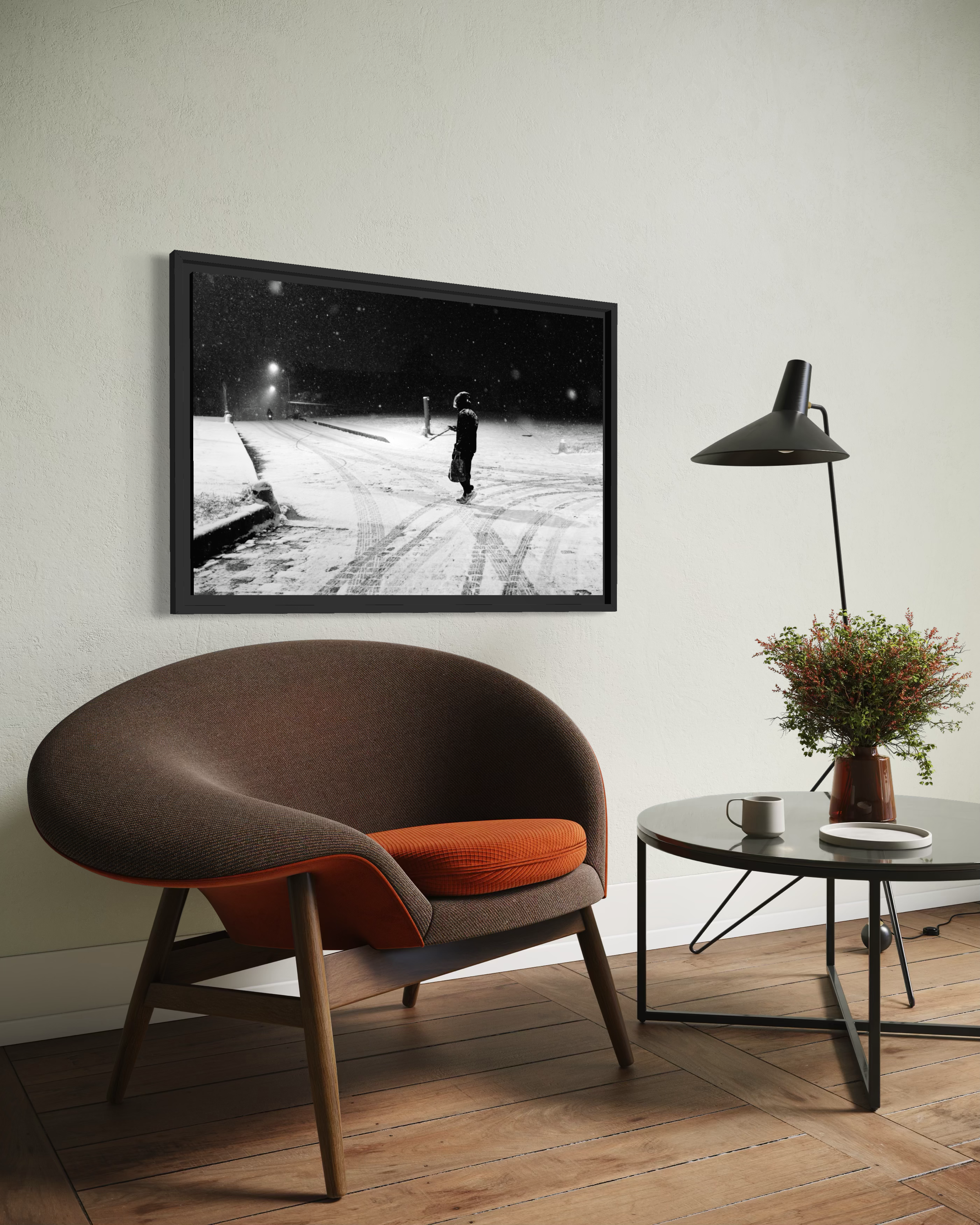 Une pièce intérieure avec un fauteuil en bois et tissu marron avec un coussin orange, une table ronde noire avec une tasse blanche et un plateau blanc, une plante verte dans un vase en verre brun, un lampadaire noir, un tableau noir et blanc représentant une scène de neige avec une personne, sur un mur blanc.