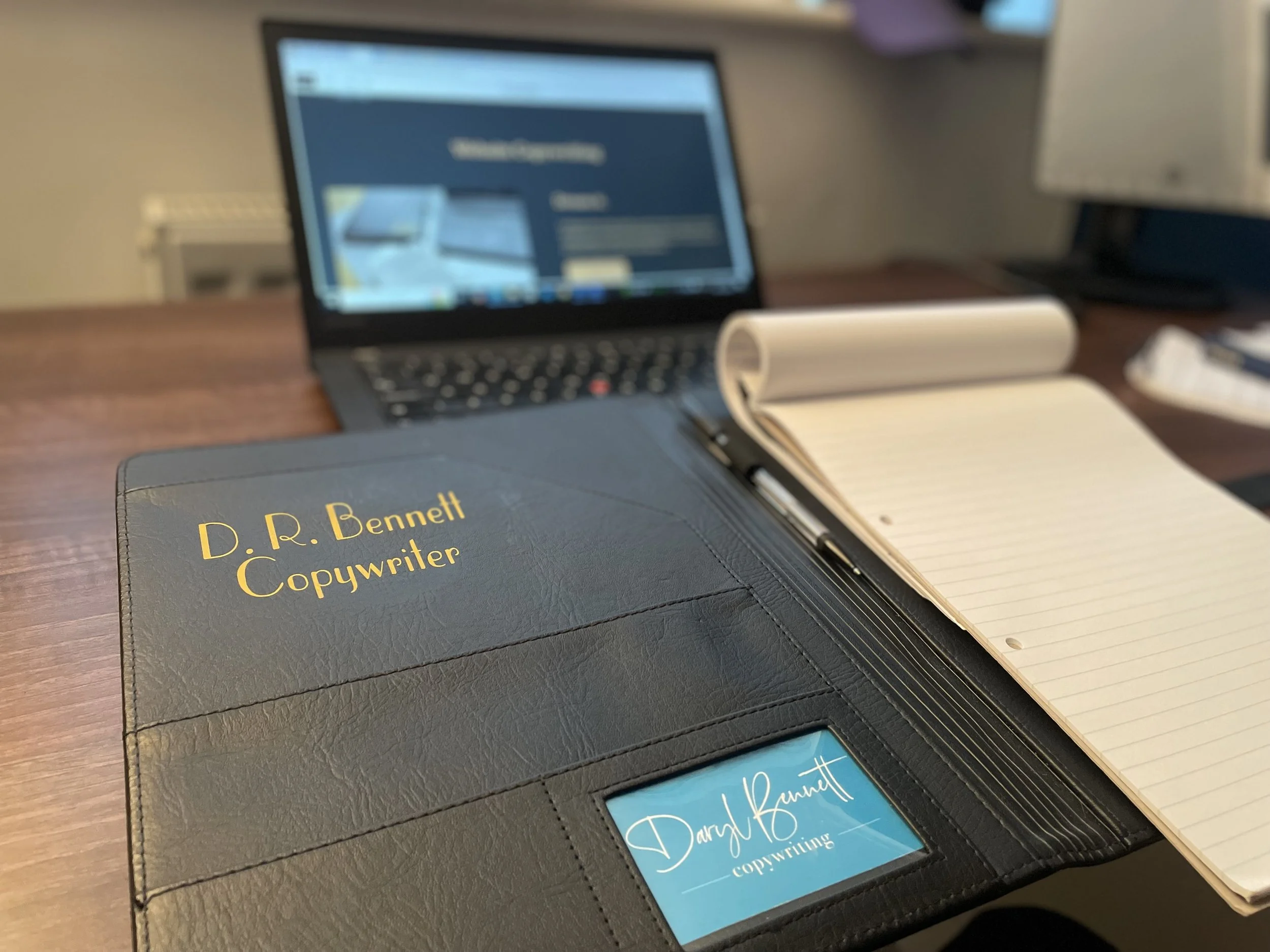 A partially open black leather planner with gold embossed text that reads "D. R. Bennett copywriter" is on a wooden desk. Next to it is a spiral notebook with handwritten notes. In the background, there is a laptop with a slightly blurred screen displaying a website or document, and a white computer keyboard is visible.