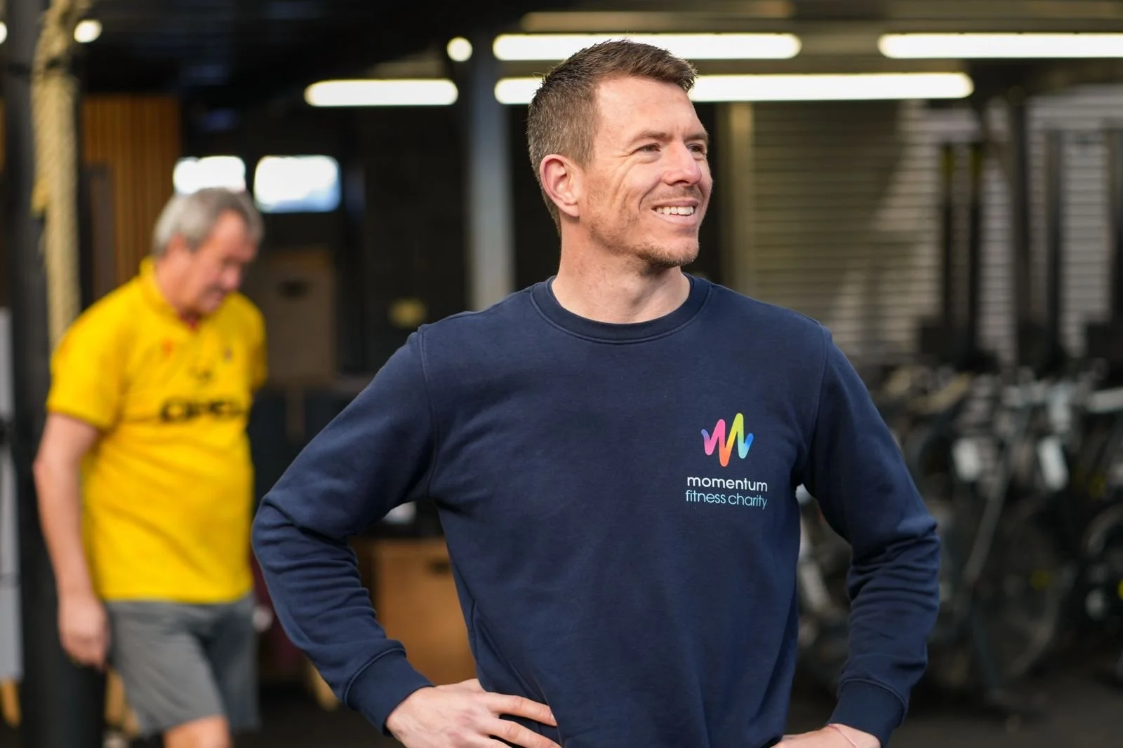 A man smiling wearing a blue sweatshirt with a colorful logo and the words 'momentum fitness charity' in a gym, with another man in a yellow shirt blurred in the background.