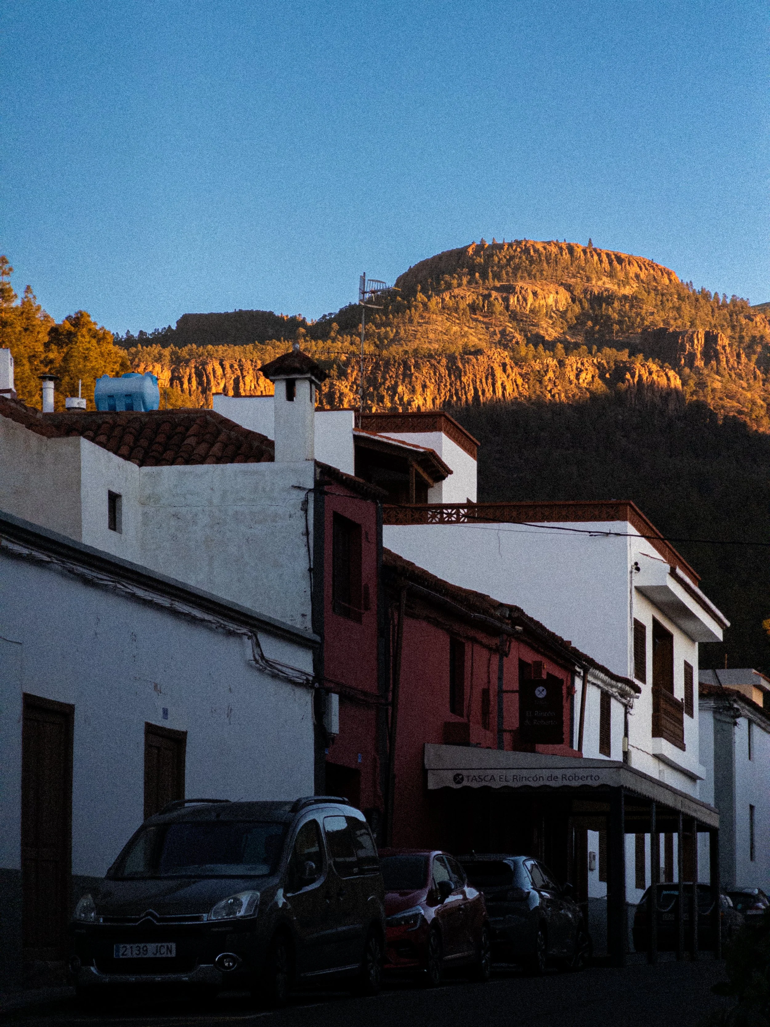 Street scene with parked cars in front of white and red buildings, with a mountain in the background illuminated by the setting sun.