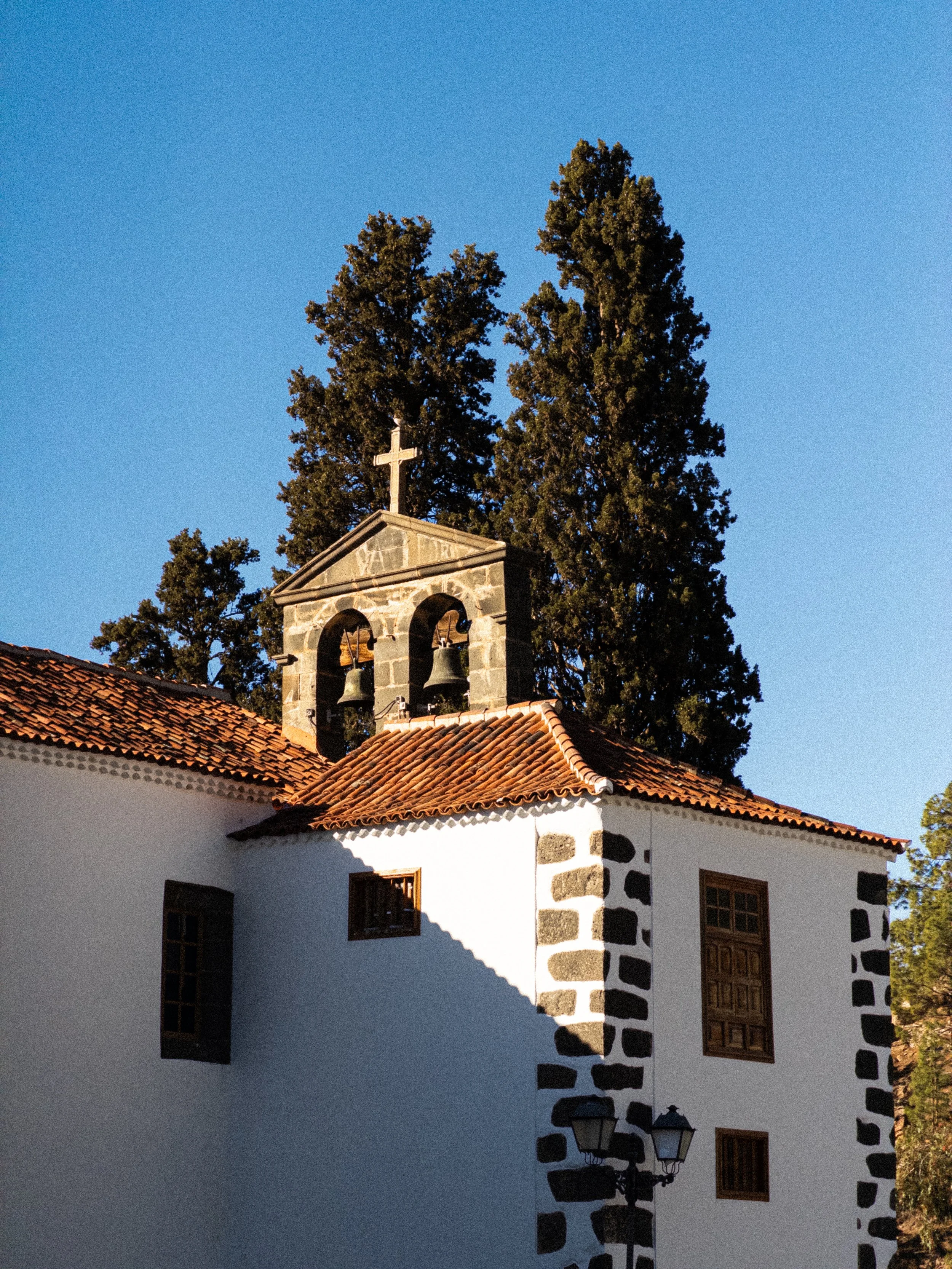 A small white church with a red-tiled roof, a stone bell tower with three bells, and a cross on top, surrounded by tall trees under a clear blue sky.