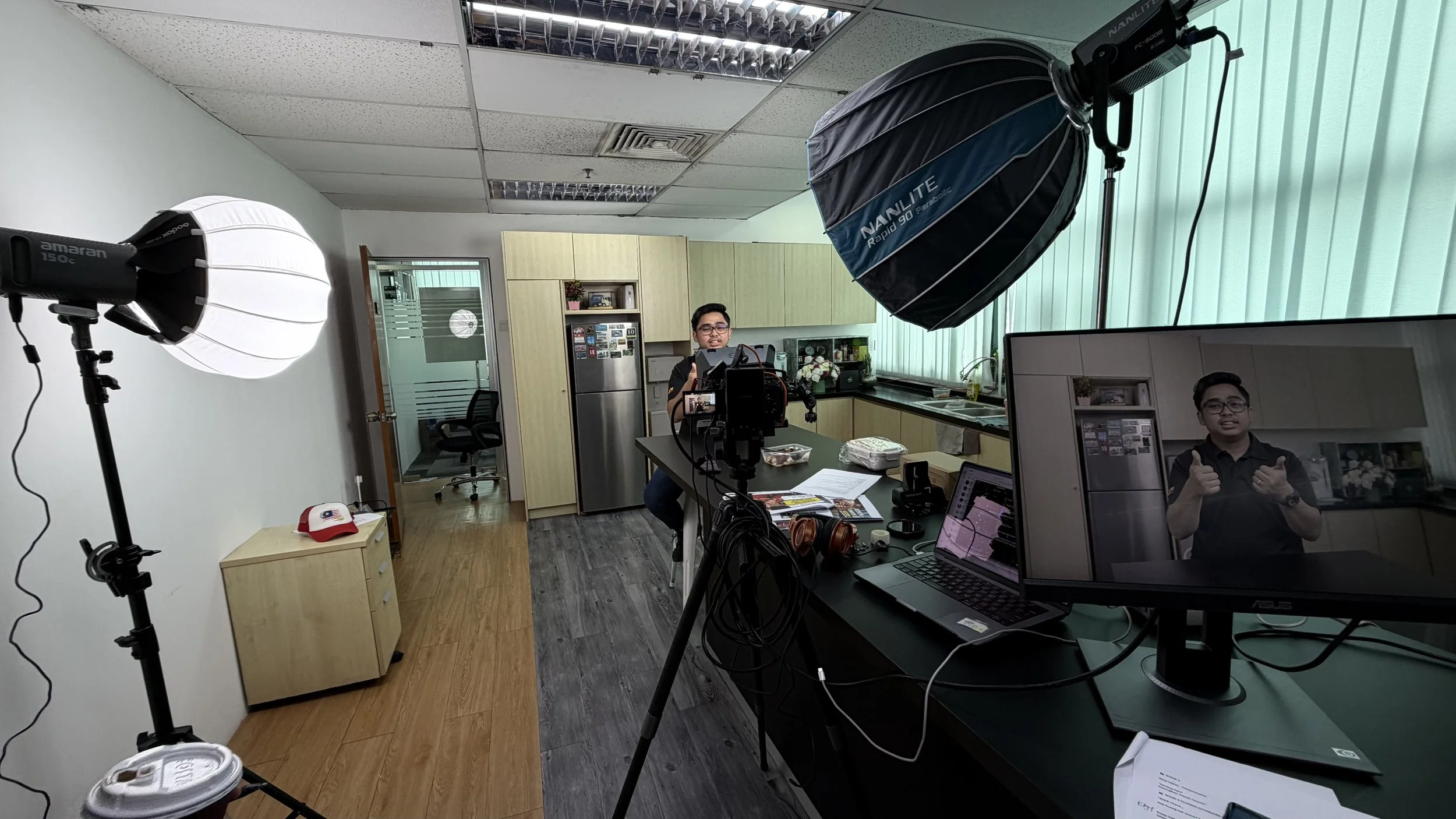 Indoor filming studio with professional lighting equipment, including two large softbox lights illuminating a man sitting at a desk and giving a thumbs-up. A camera on a tripod captures the scene, and monitors display the man. The room has a kitchen area with a refrigerator, shelves, and decorative items, with a window covered by vertical blinds.