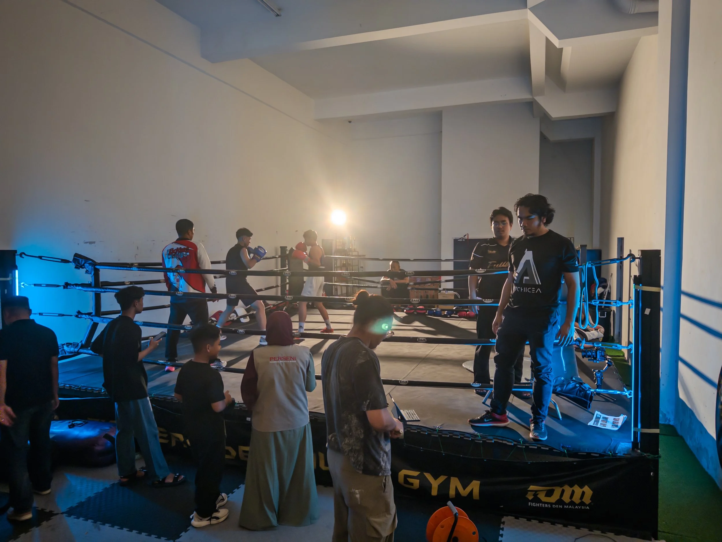 A group of people participating in a boxing training session inside a gym. Some are in the ring, wearing boxing gloves, while others are standing or sitting around the ring observing. There is a bright light source in the background illuminating the scene.