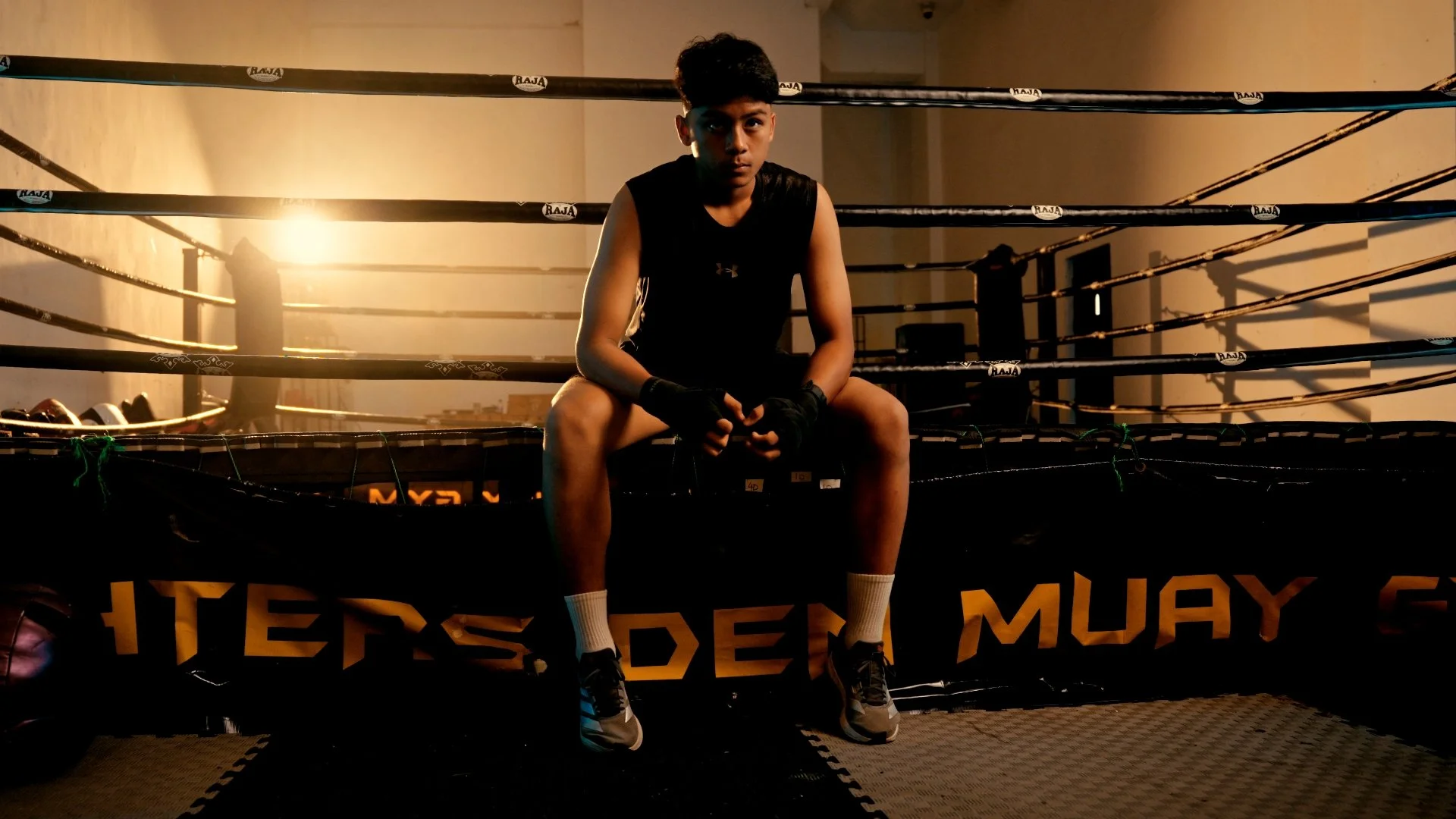 A young male boxer sitting in a boxing ring with sunlight shining behind him, wearing sports attire including gloves, shorts, and shoes, in a dimly lit gym.
