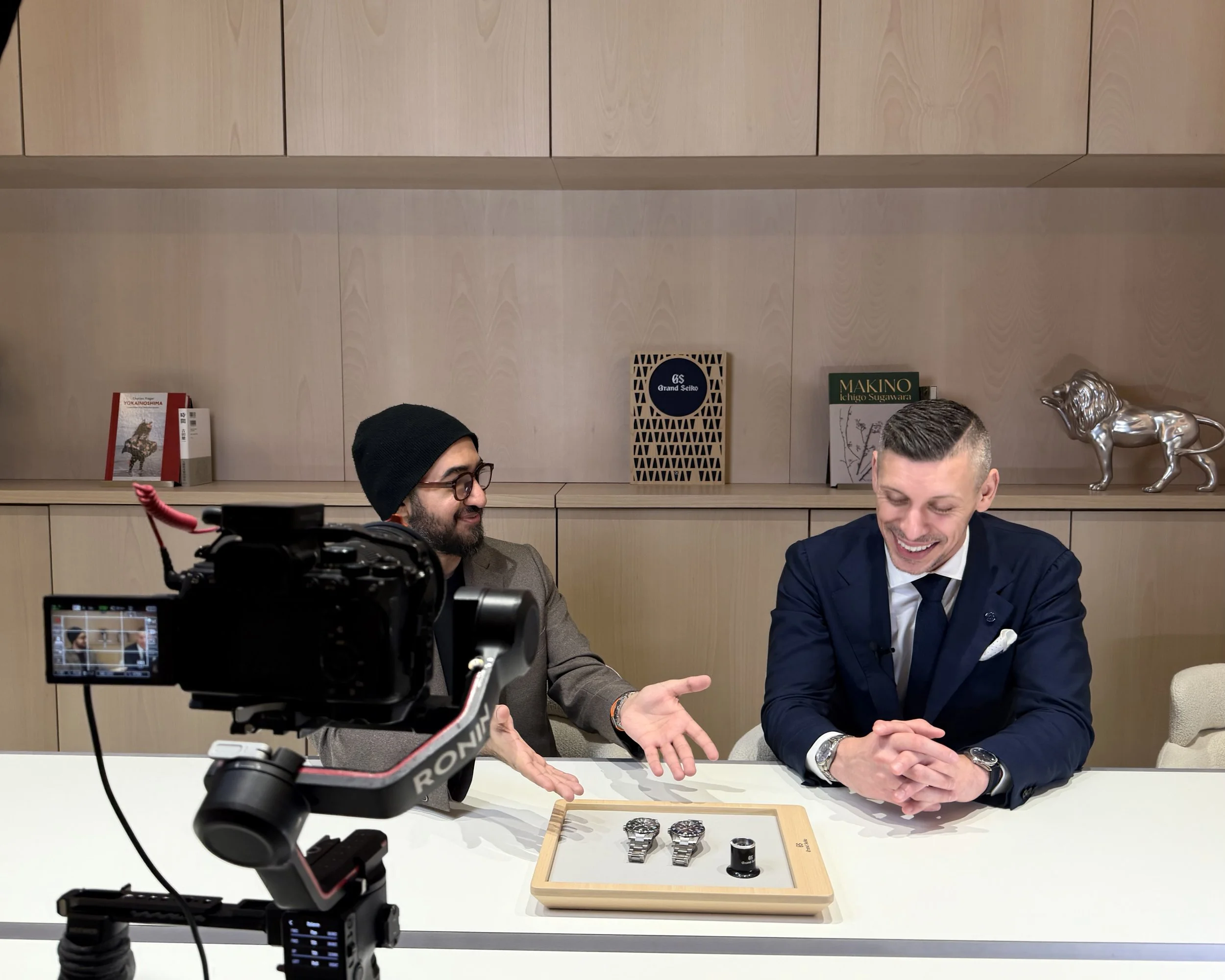 Two men are sitting at a table with watches and jewelry displayed on a tray, engaging in a conversation. One man is wearing a dark suit, and the other is wearing glasses and a beanie. A camera is set up in front of them, recording the interaction.