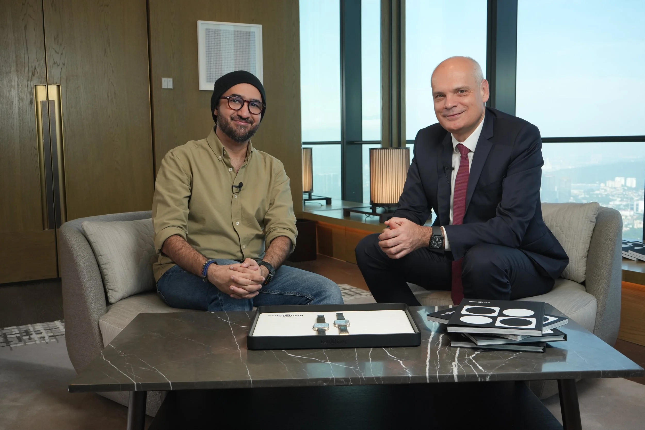 Two men sitting on a sofa in a modern office with large windows. One man is wearing a beige shirt, glasses, and a black beanie; the other is in a dark suit with a white shirt and a red tie. A coffee table with electronic devices and papers is in front of them.