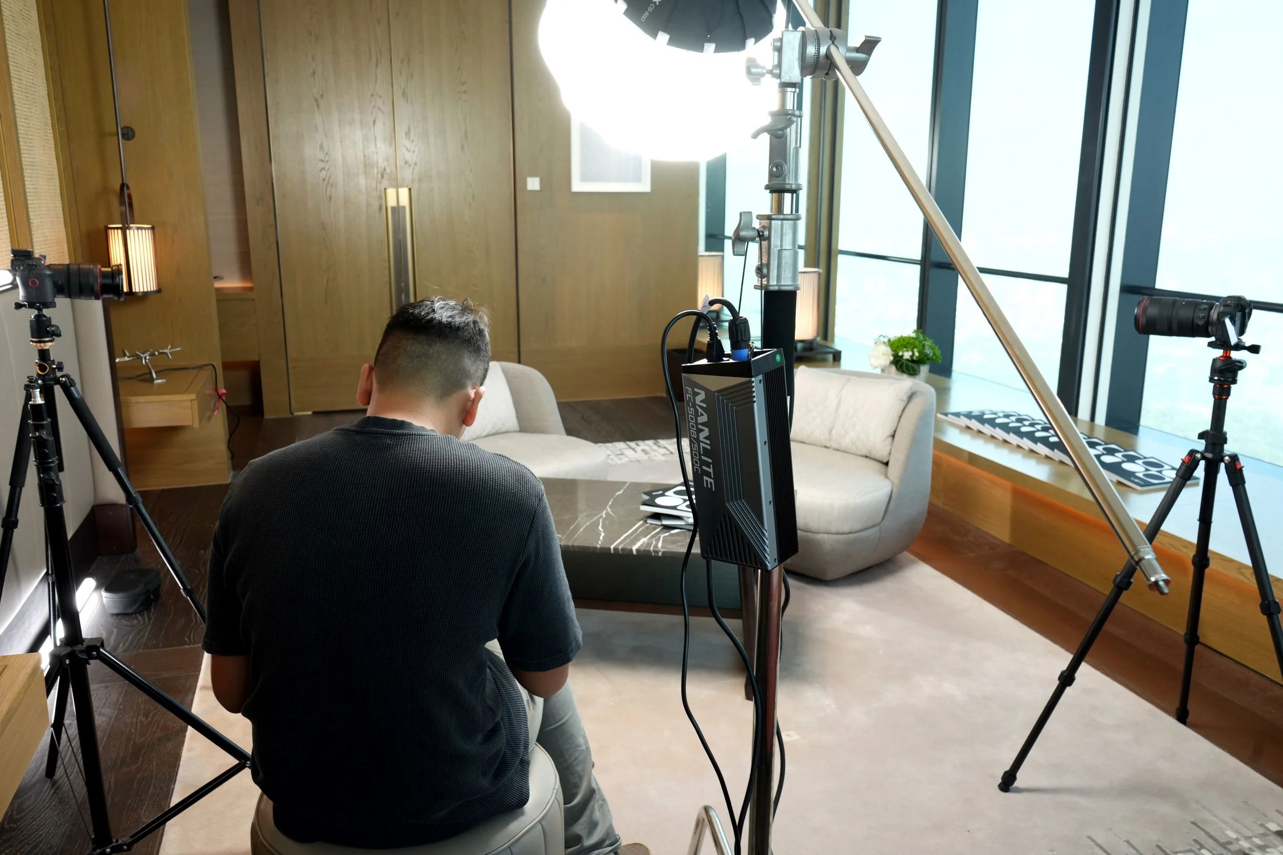 An indoor setting with a man sitting on a stool, facing away, surrounded by photography equipment including tripods with lights and cameras, in front of a large window with natural light, a couch, a small table, and decorative elements such as a potted plant and lamps.