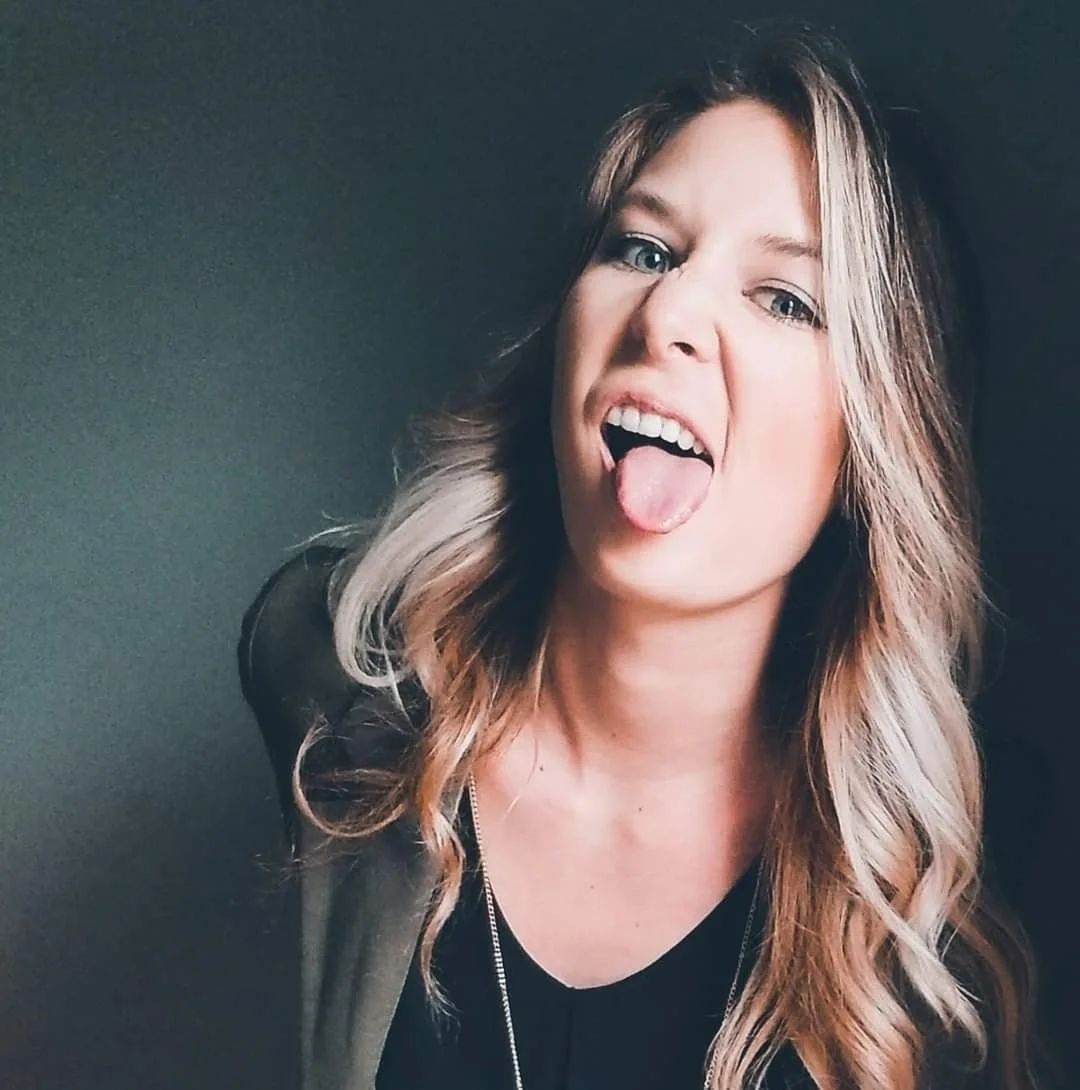 A woman with long wavy hair making a silly face, sticking out her tongue, against a plain dark background.
