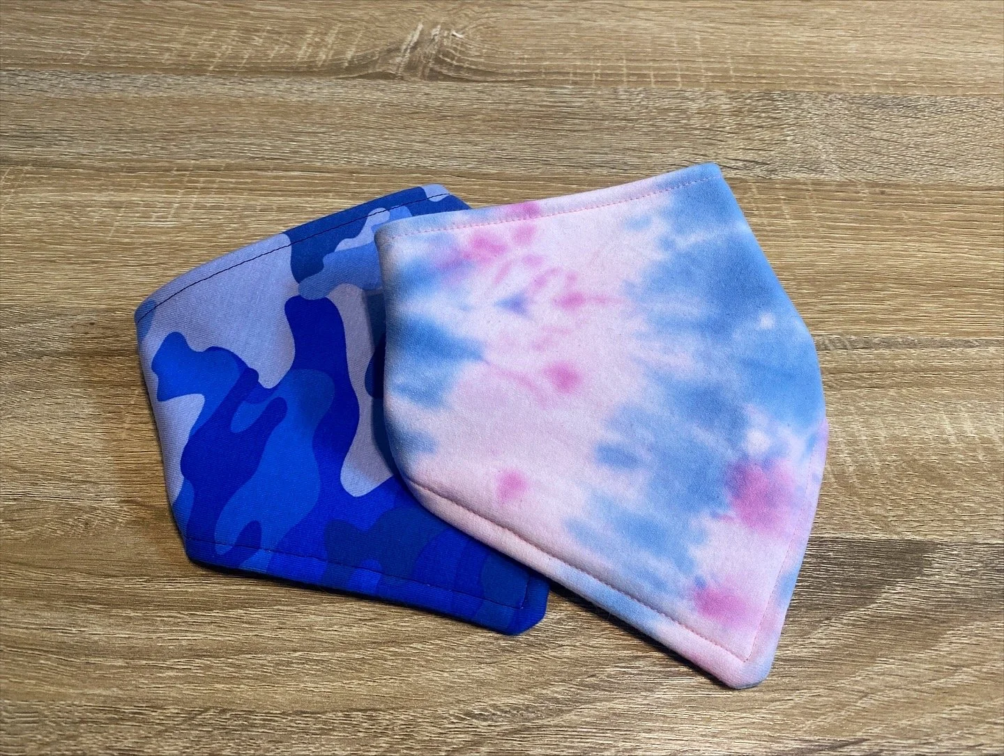Cooling dog bandanas displayed on wood background, featuring a blue camouflage design and a pink and blue tie-dye print, handmade pet accessories by The Fluffed Tail.