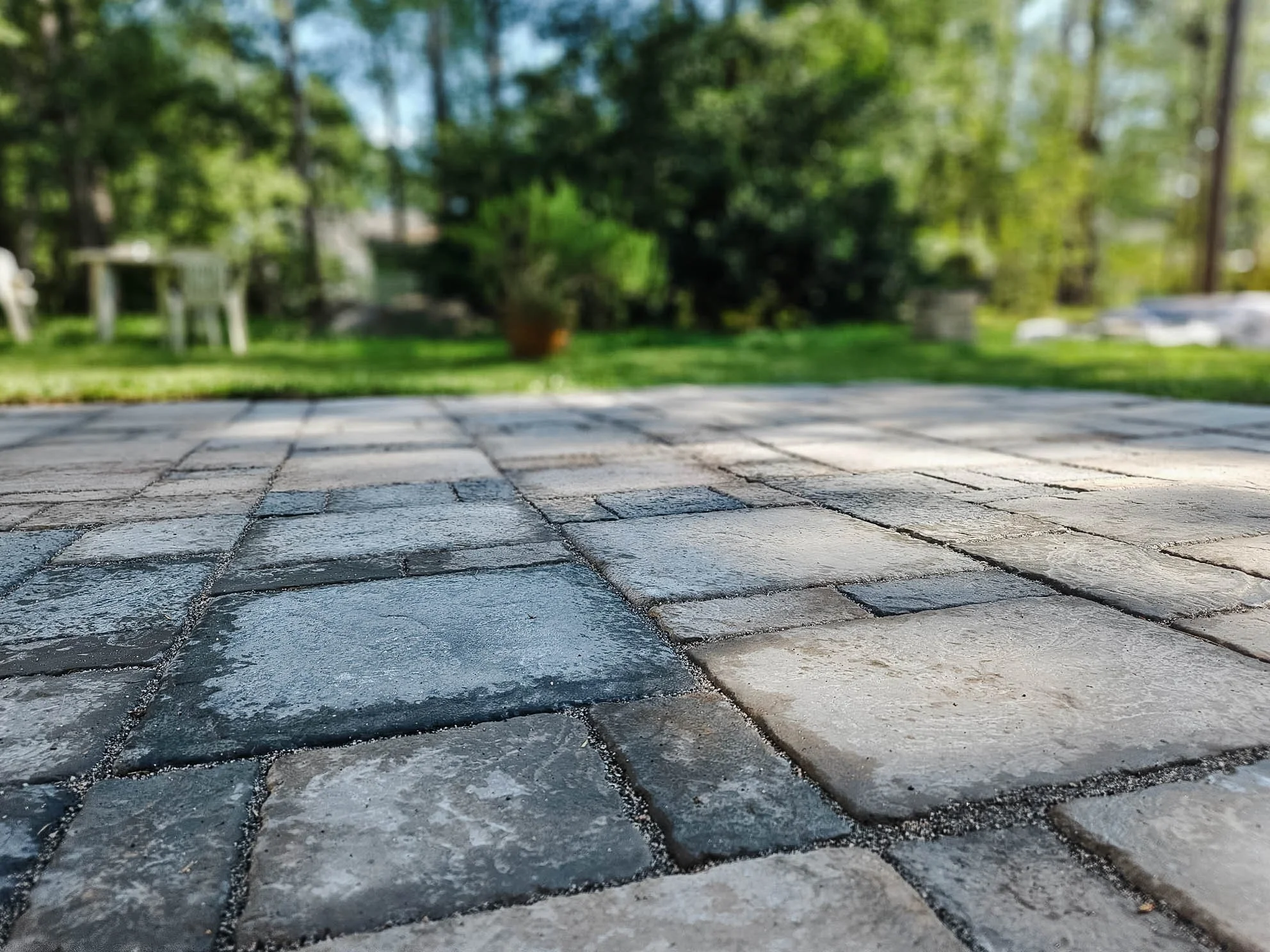 Close-up view of a paved stone patio with various gray and beige stones, with a blurred green backyard in the background.