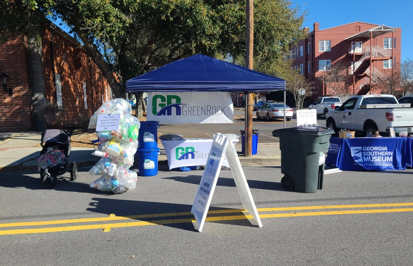 A street-side event booth with a blue canopy featuring the Greenrock logo, a table with a blue tablecloth for Georgia Southern Museum, recycling bins, a signboard, a bag of empty plastic bottles, and a stroller, with parked cars and a brick building in the background.