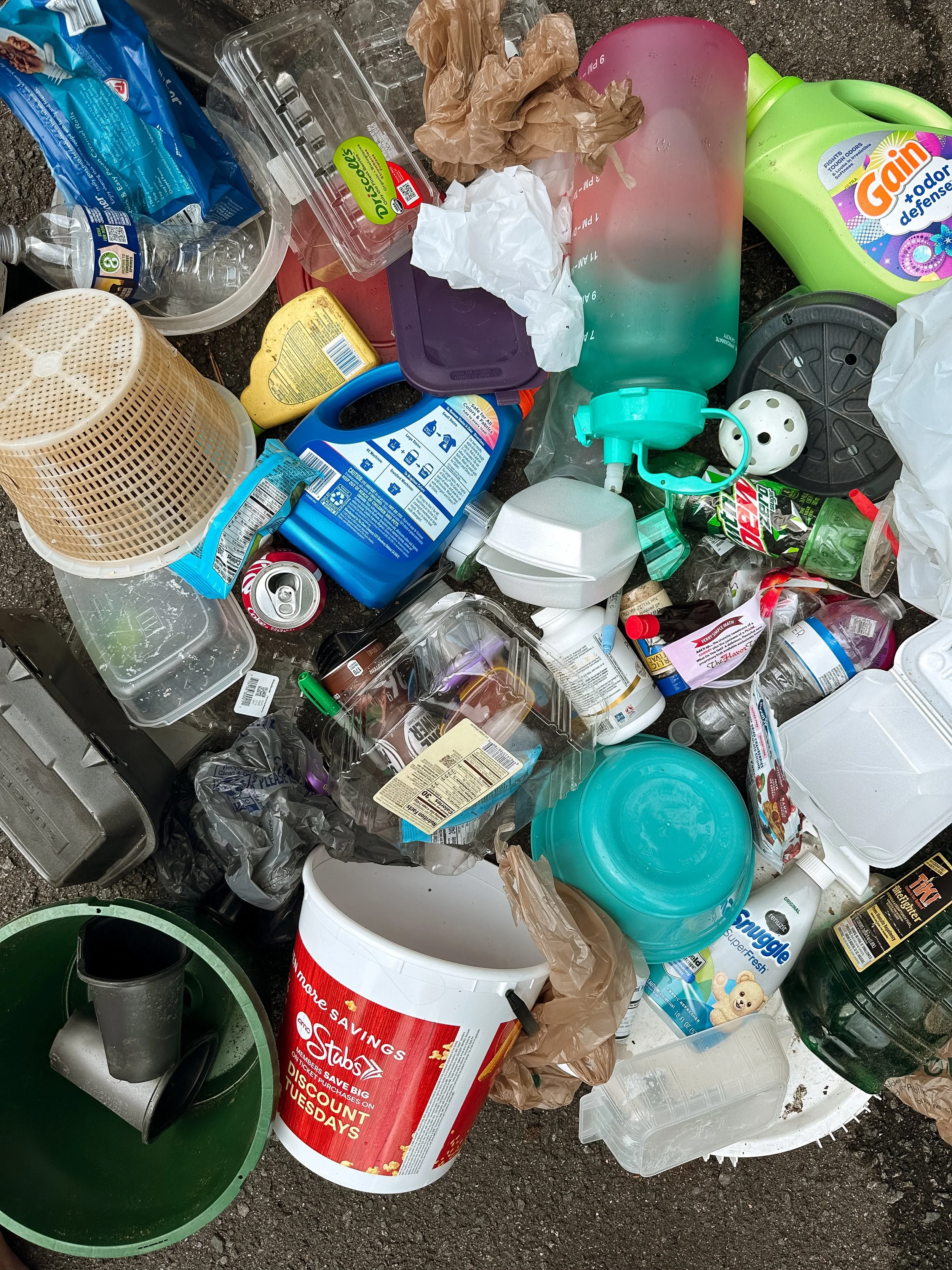 Mixed trash and recyclables on the ground including plastic bottles, containers, paper, and a bucket.