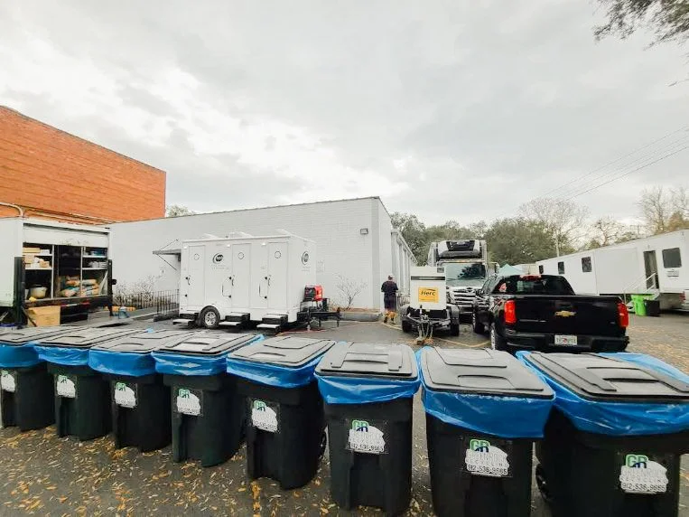 Rooftop food truck in a parking lot with a row of black trash bins with blue lids in the foreground, several trailers and trucks, one with a yellow 'FIRE' sign, and a person standing near the trailers, under a cloudy sky.