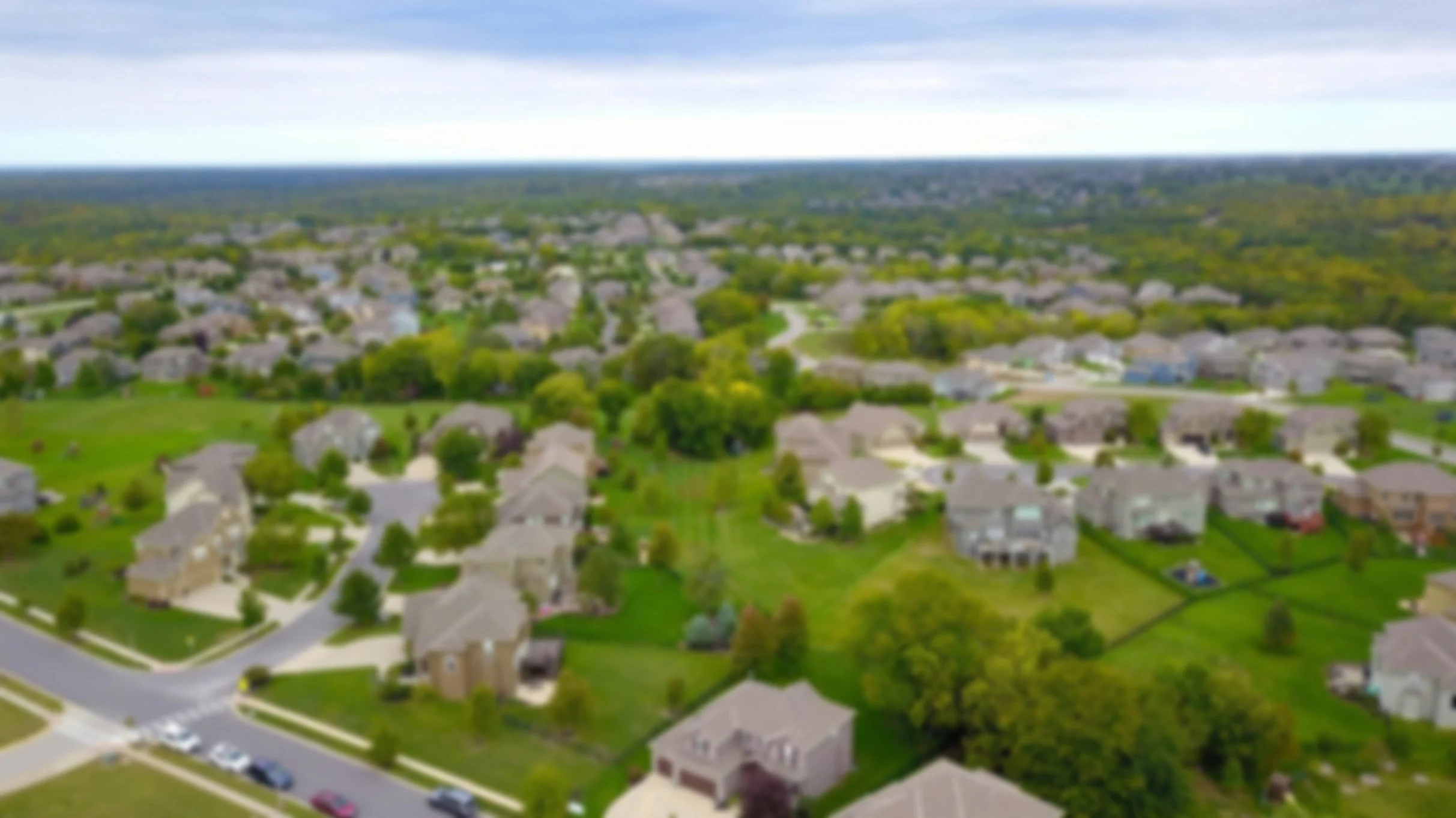Aerial view of a suburban neighborhood with houses, trees, and green lawns under a partly cloudy sky.