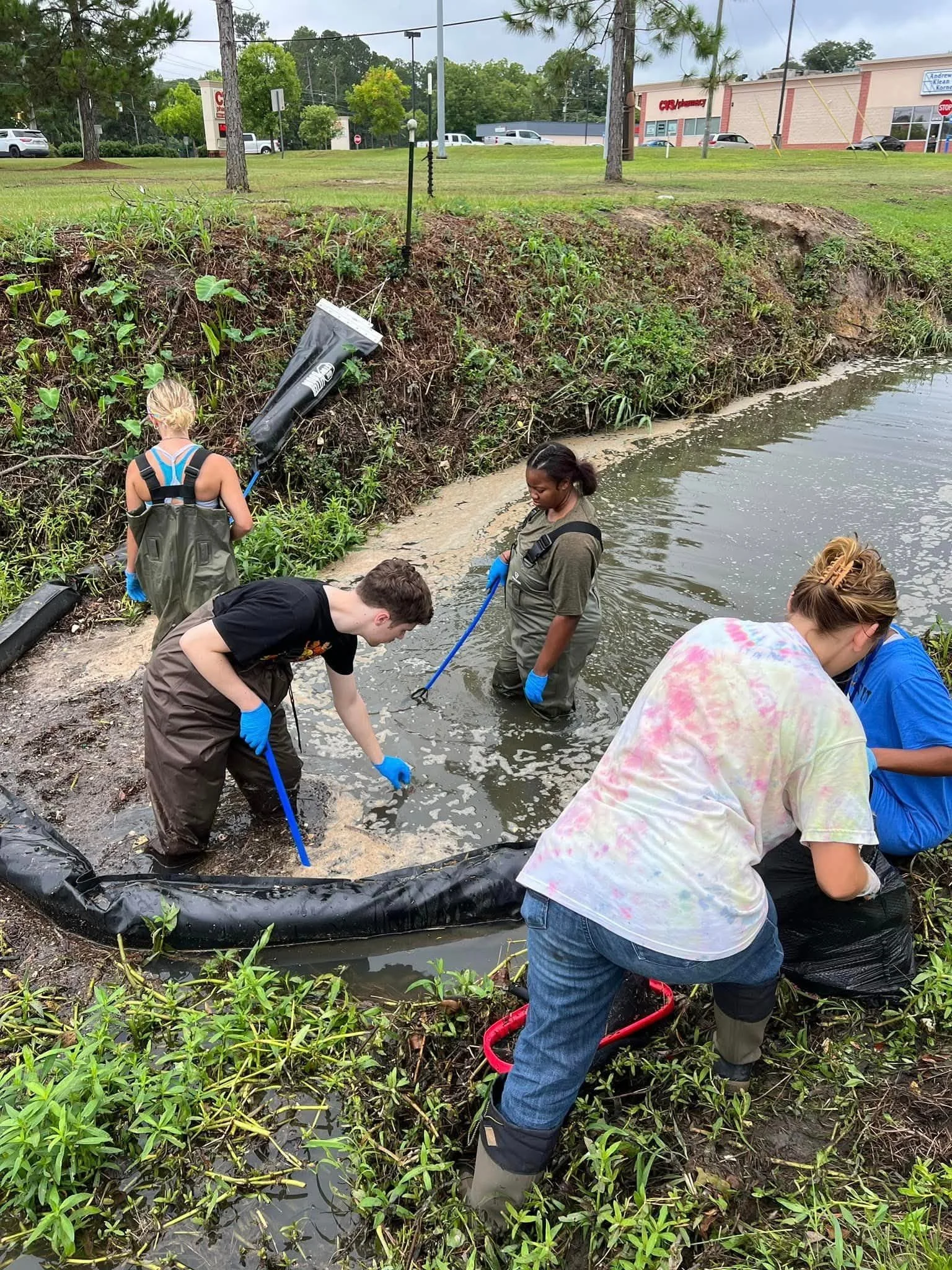 A group of people, wearing waders and gloves, cleaning trash from a pond or small lake near a grassy area with trees and buildings in the background.