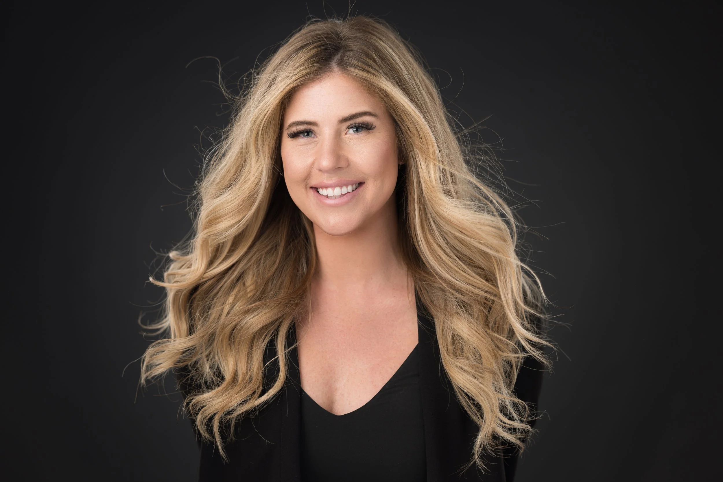 A woman with long blonde hair is sitting on outdoor metal stairs, smiling and looking at the camera. She is wearing a tan blazer, brown top, blue jeans, and tan platform sandals. The background includes a brick wall and windows.