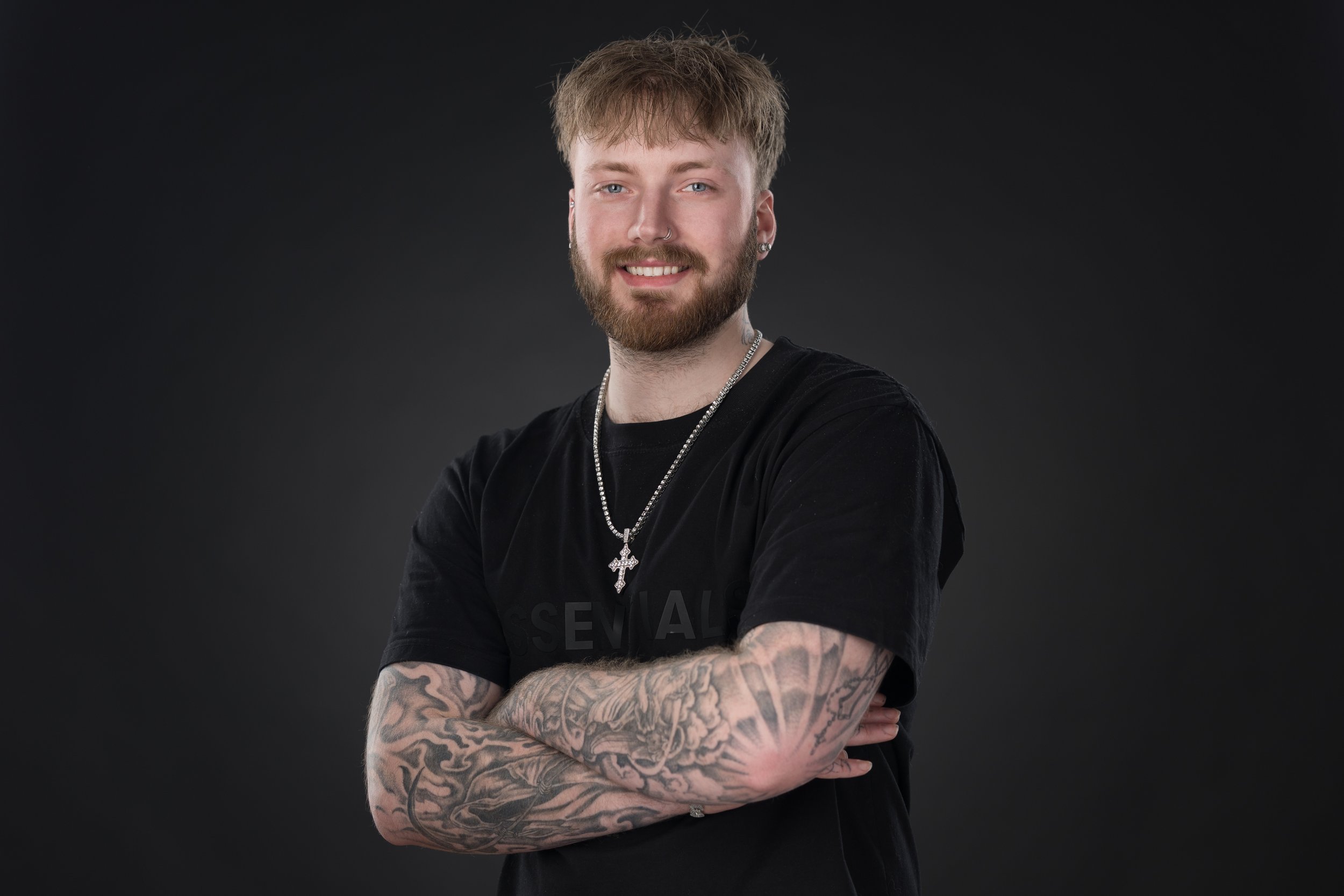 A young man with blond hair, a beard, and tattoos on his arms, smiling and leaning against a brick wall, wearing a black NASCAR t-shirt and beige shorts.