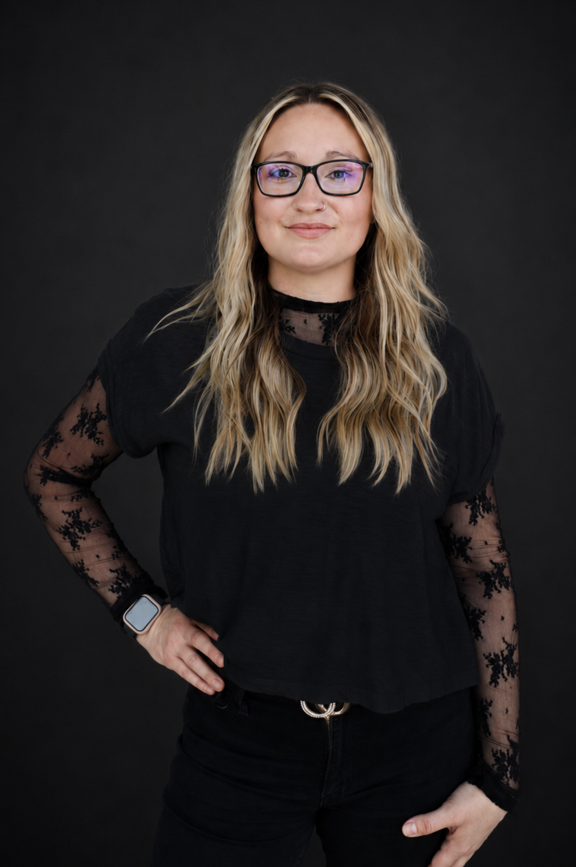 Young woman with long blonde wavy hair, glasses, and a nose piercing, wearing a white top with black lace long sleeves, standing against a brick wall.