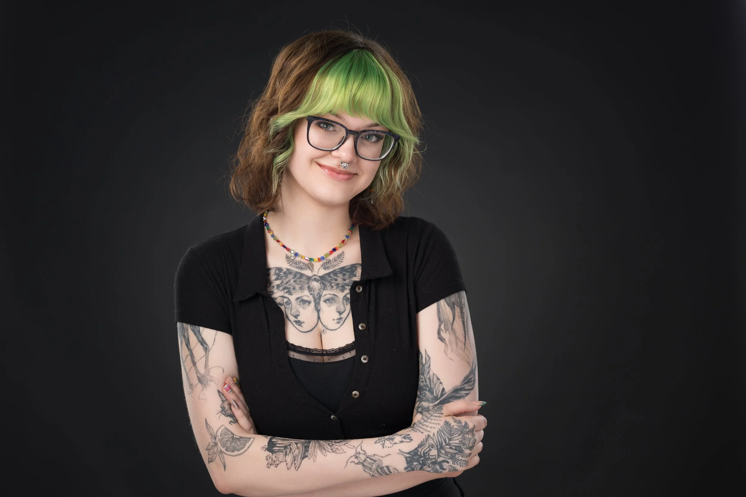 A woman with dyed green hair, wearing glasses, a black off-shoulder top with floral patterns, black pants, and black high-top sneakers, sitting on metal stairs outdoors near a brick building.
