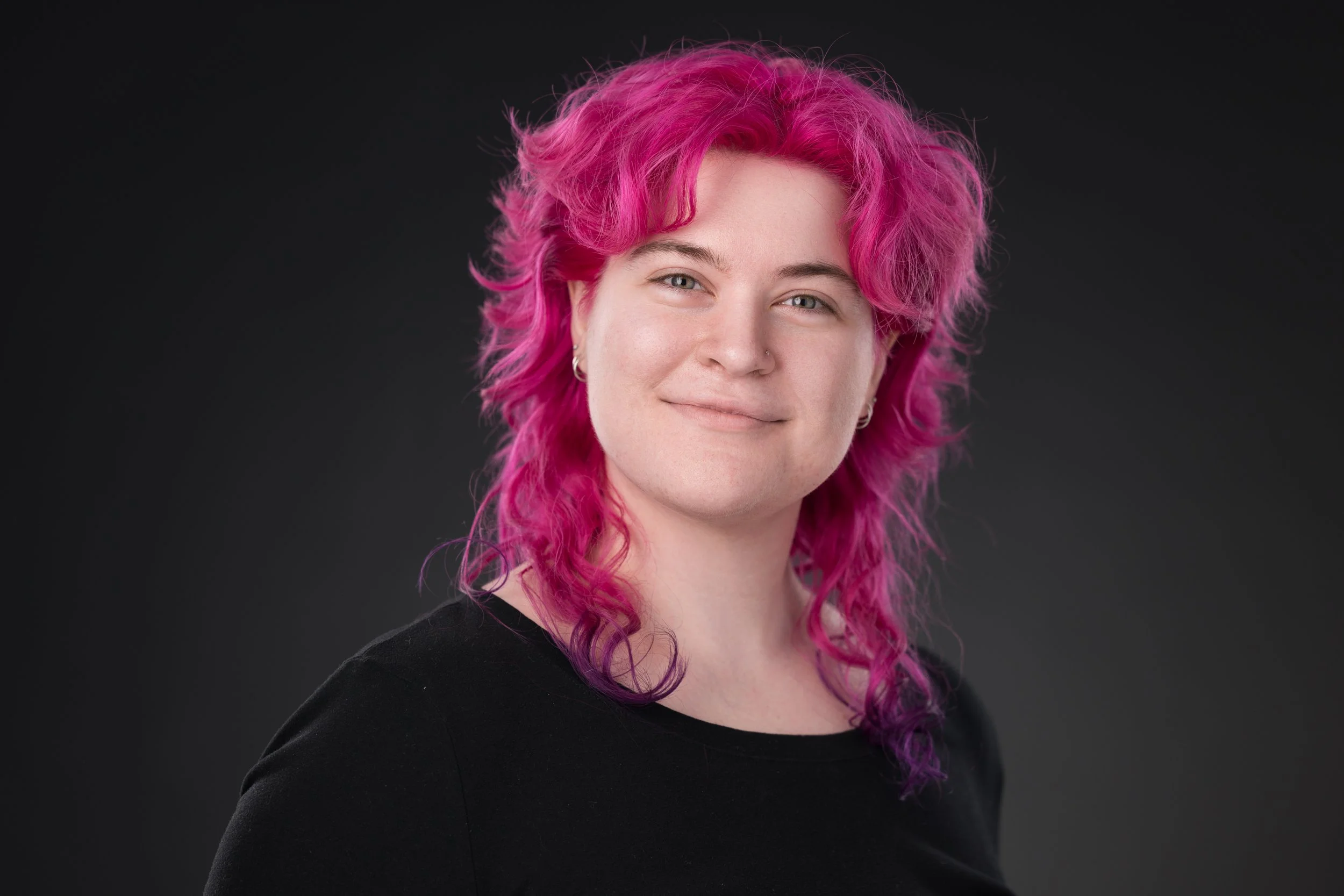 A young woman with vibrant pink and purple hair, sitting on metal stairs outside near a brick wall, smiling at the camera.