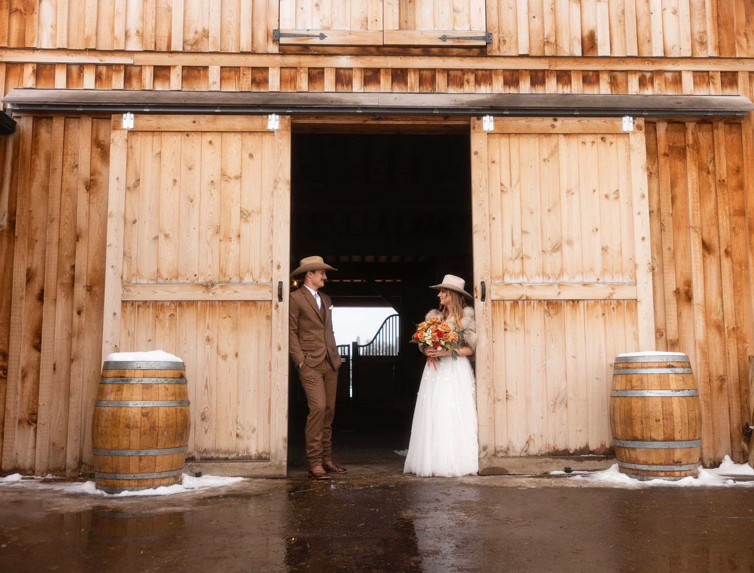 A couple wearing western wedding attire pose in a rustic barn.