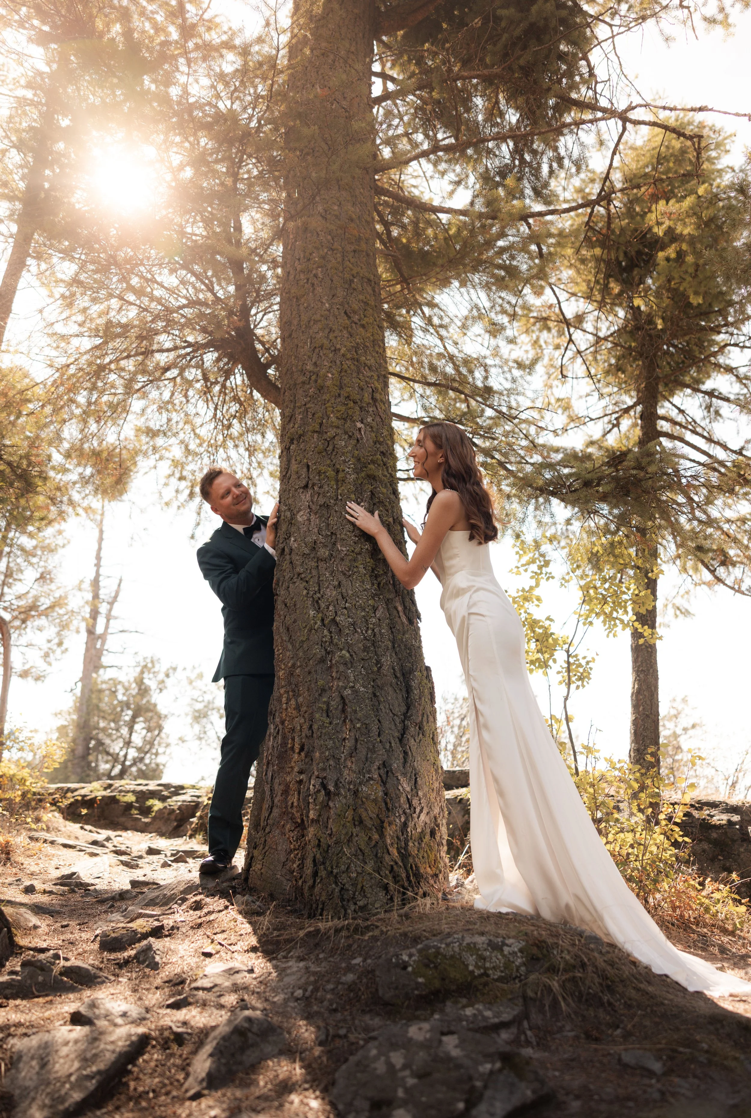 A bride and groom play peek-a-boo around a tree in a stunning forest setting.