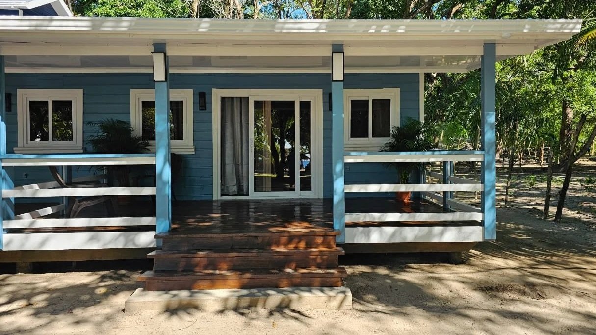 A small blue wooden cabin with a front porch surrounded by trees, featuring white framed windows and a glass door.
