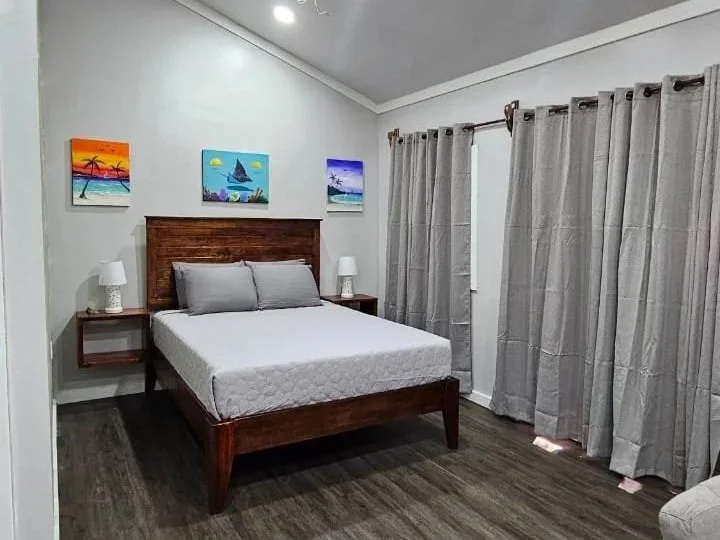 Modern bedroom with a wooden bed frame, gray bedding, two nightstands with lamps, abstract paintings on the wall, gray curtains, and wooden flooring.