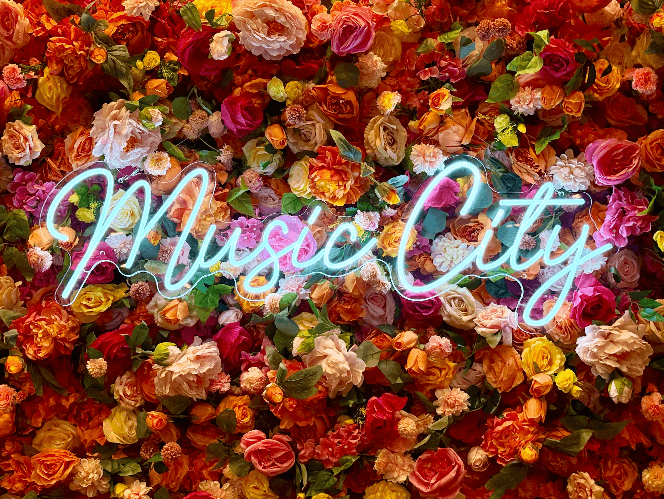 A bright white Music City neon sign rental displayed on a vibrant red, orange, and pink wildflower wall backdrop.