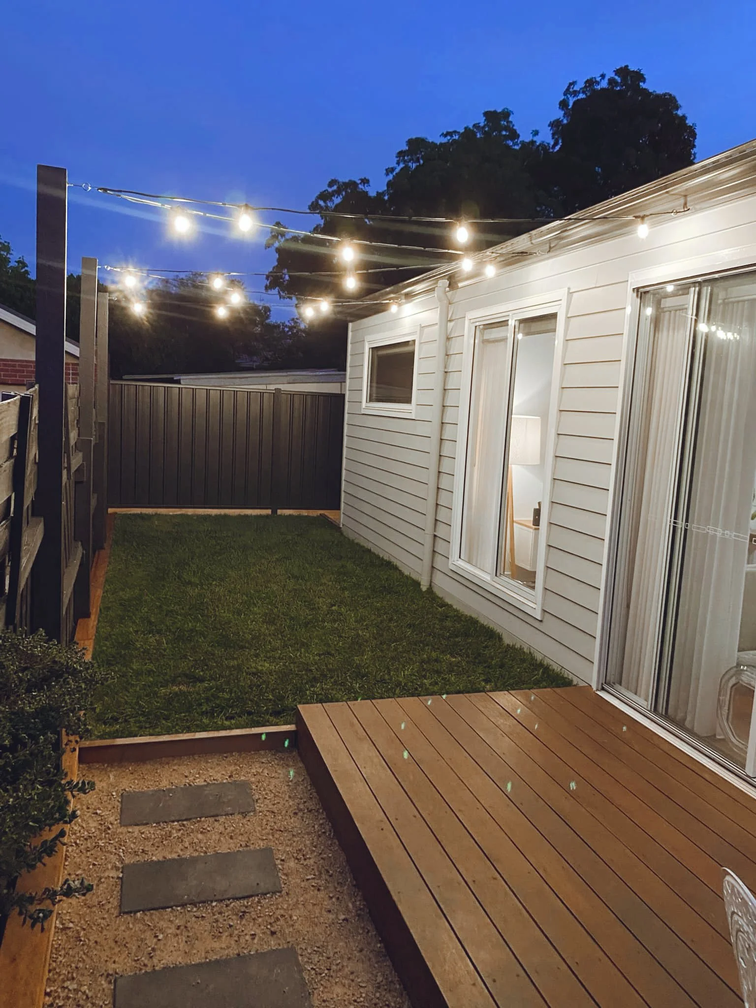 This image shows an outdoor backyard scene at dusk with string lights hanging overhead. There is a small wooden deck attached to a white house with sliding glass doors and windows. A grassy lawn is visible, with a fence and trees in the background.