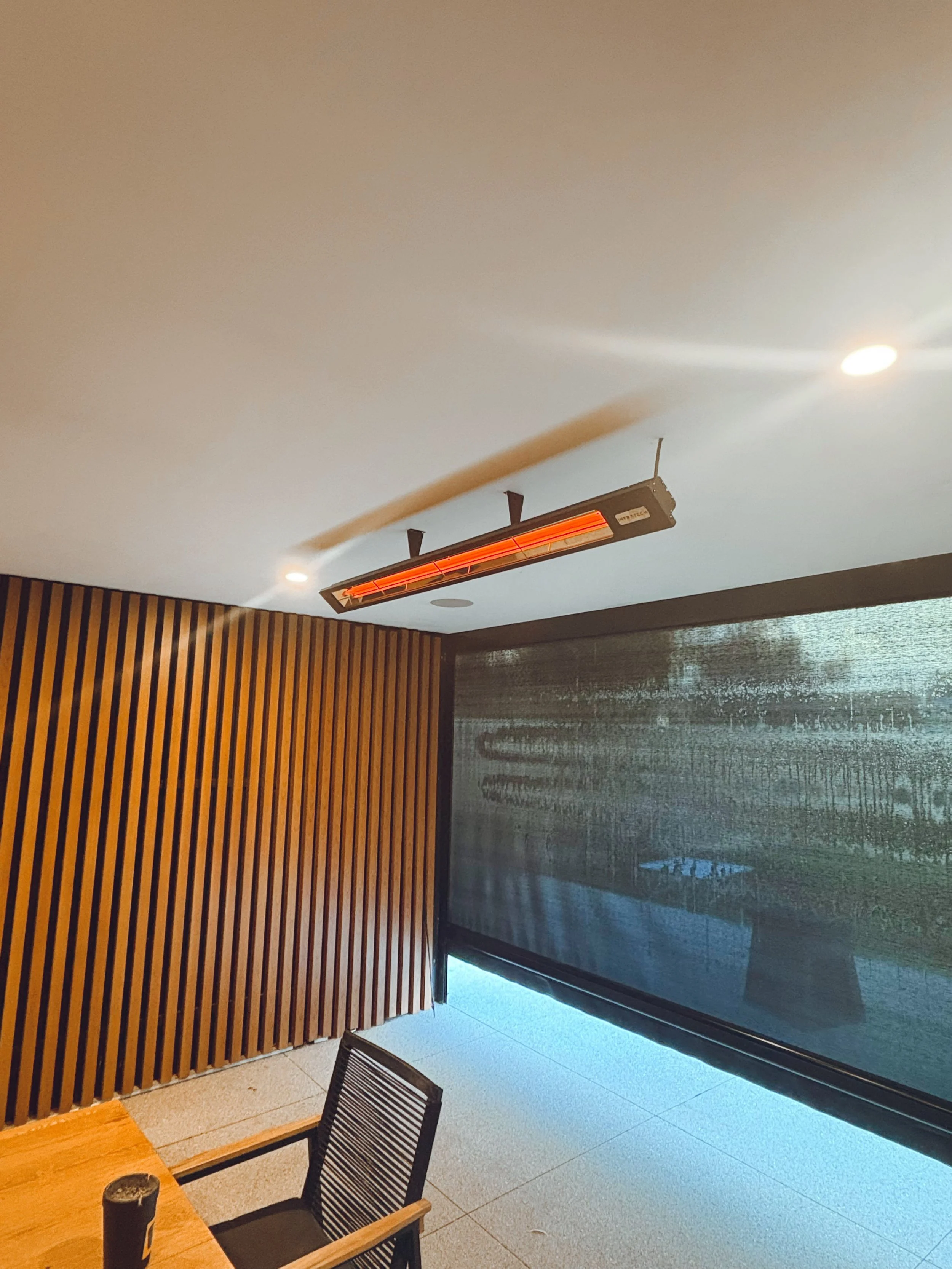 Indoor space with wooden wall panel, a chair, a table, a window with a screen, painted ceiling, and a ceiling-mounted infrared heater.