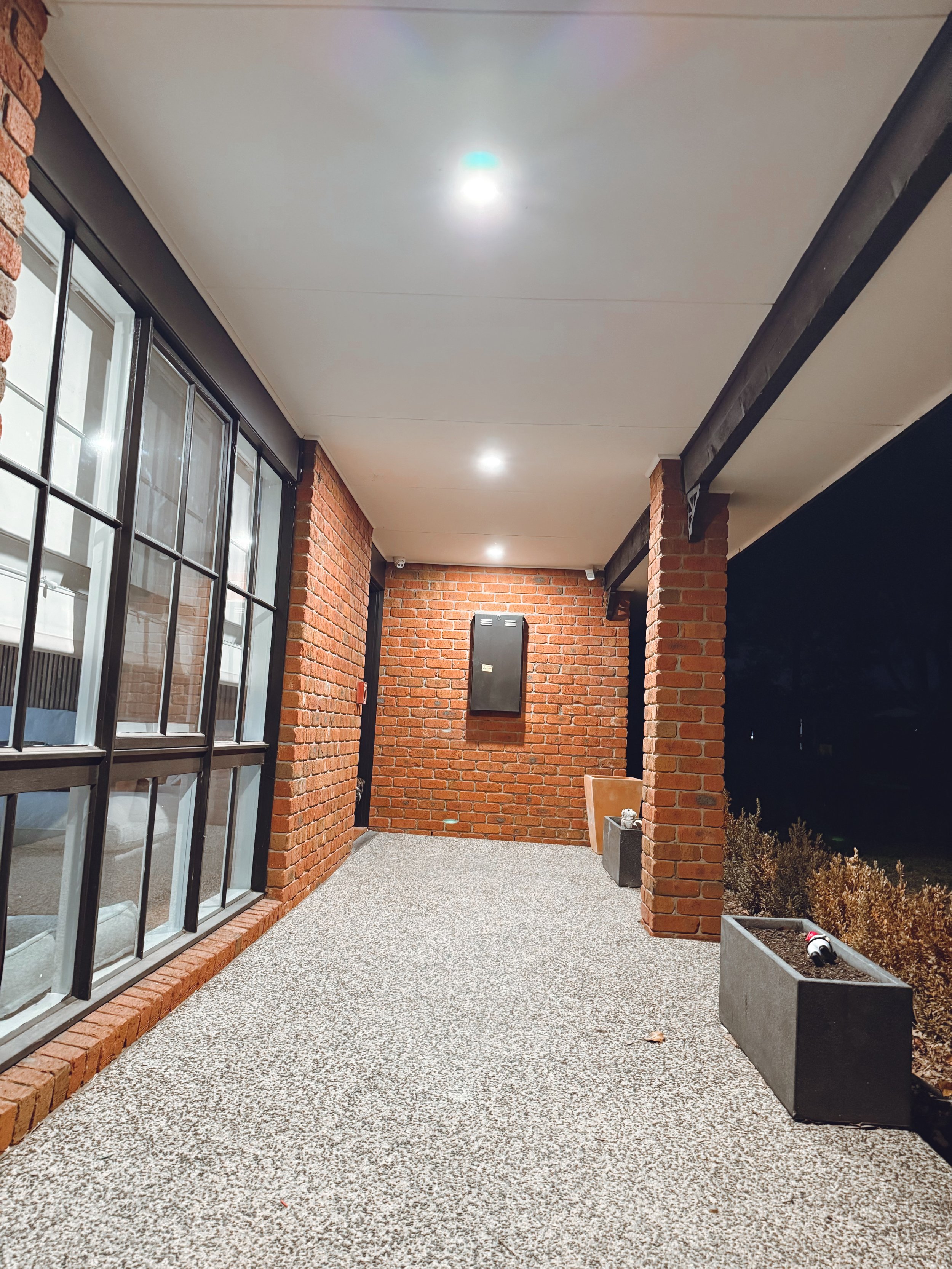 Nighttime view of a covered porch with brick walls and floor-to-ceiling windows, illuminated ceiling lights, potted plants, and a bench.
