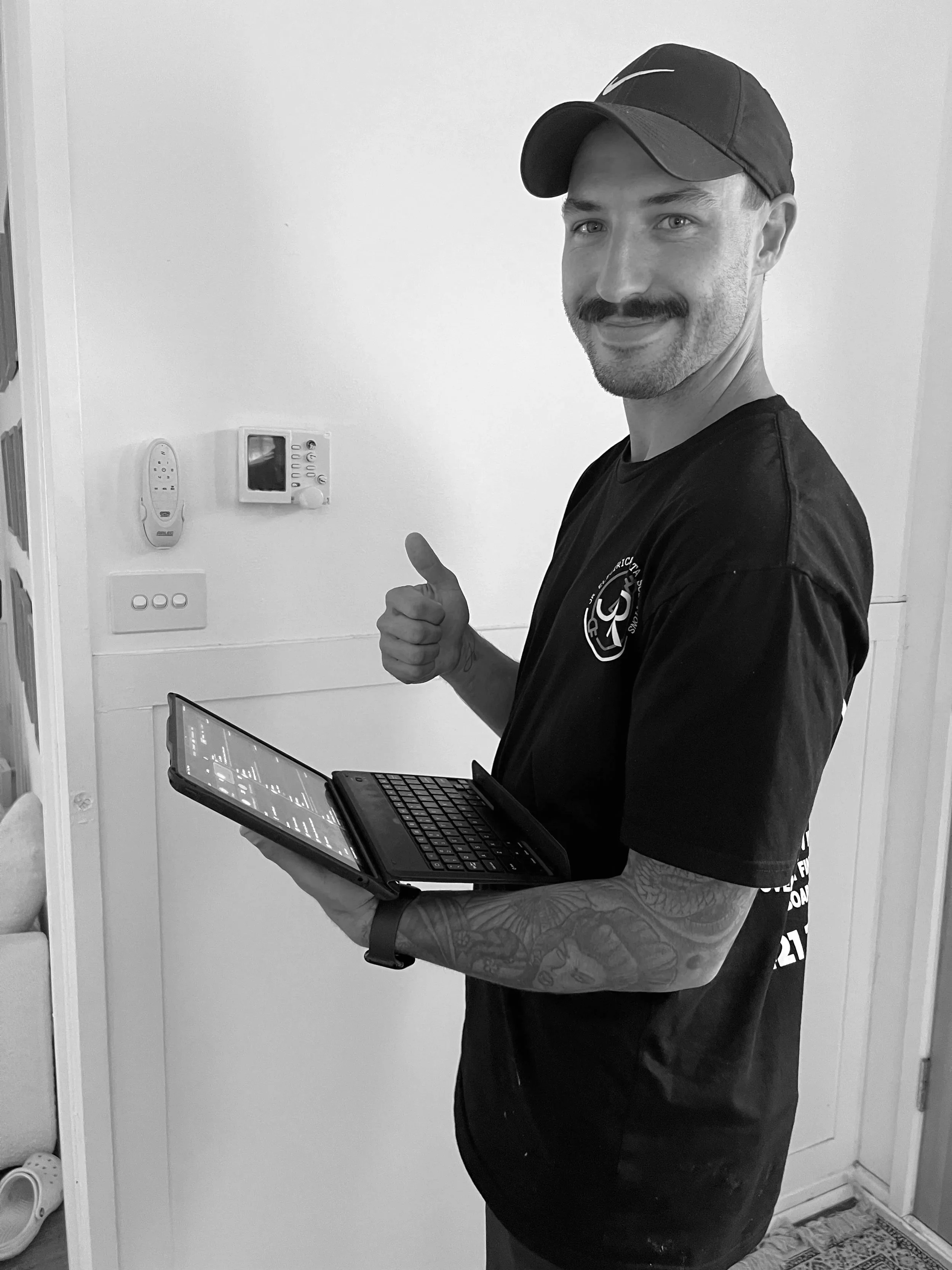 A man with a mustache and tattoos on his arm is smiling and giving a thumbs-up while holding a tablet device. He is wearing a black cap and a black t-shirt with a logo, standing near a wall with a thermostat and a remote control.